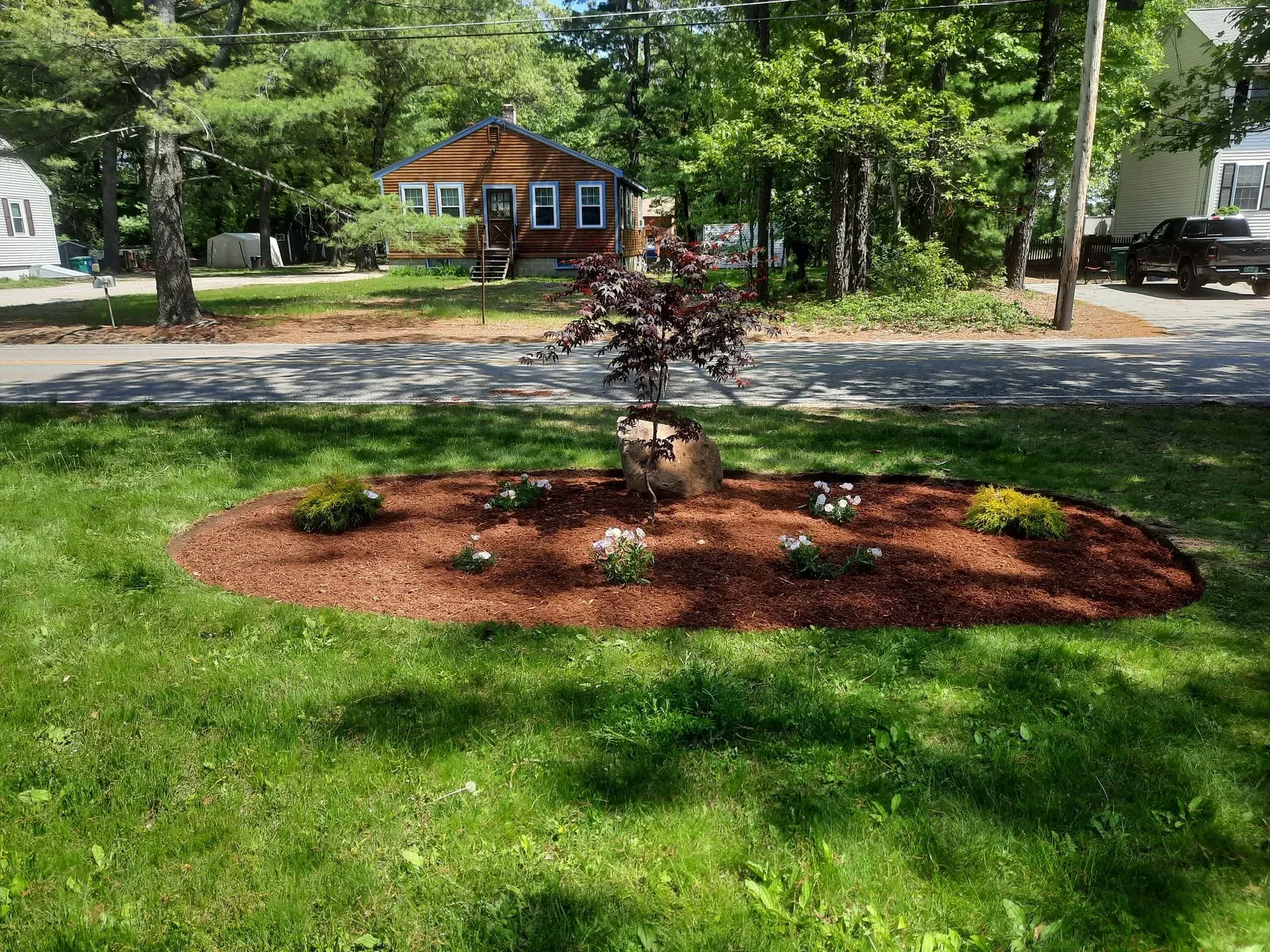 Front yard with a circular flower bed, small tree, and house across the street under green trees