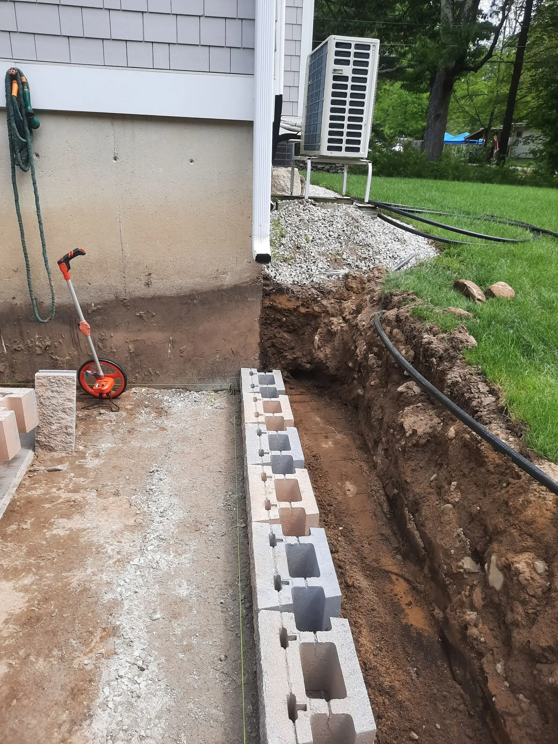 Foundation work beside a house, with cinder blocks, gravel, dirt trench, and a shovel by the wall