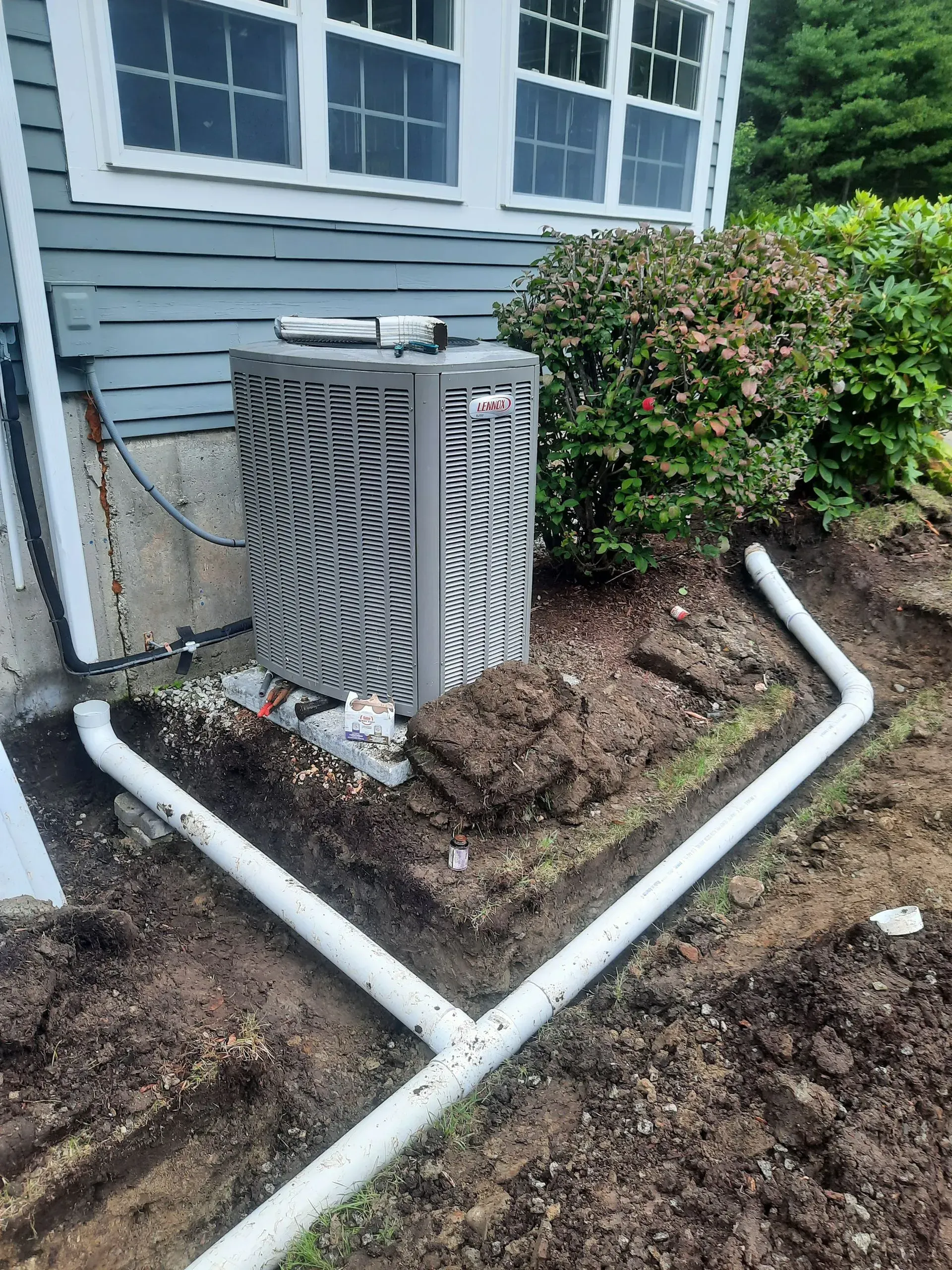 Outdoor HVAC unit beside a blue house with white pipes and bushes
