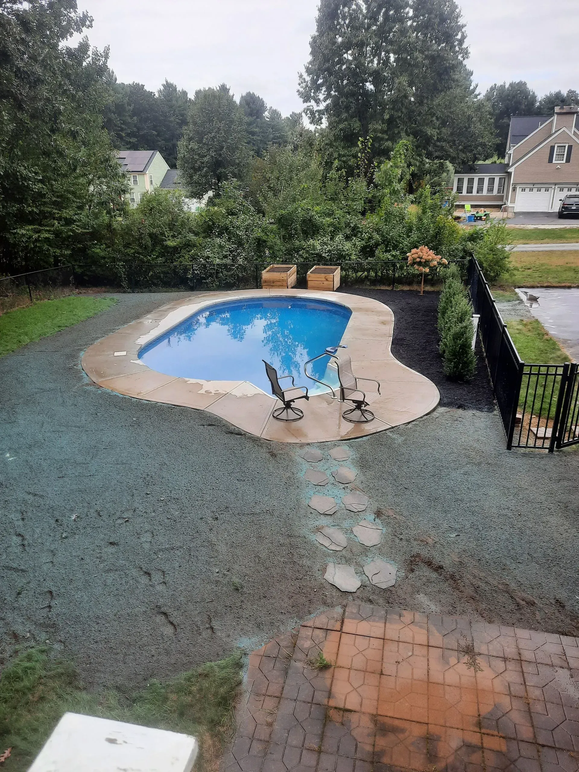 Backyard with an empty blue swimming pool, wet patio, and trees and houses in the background