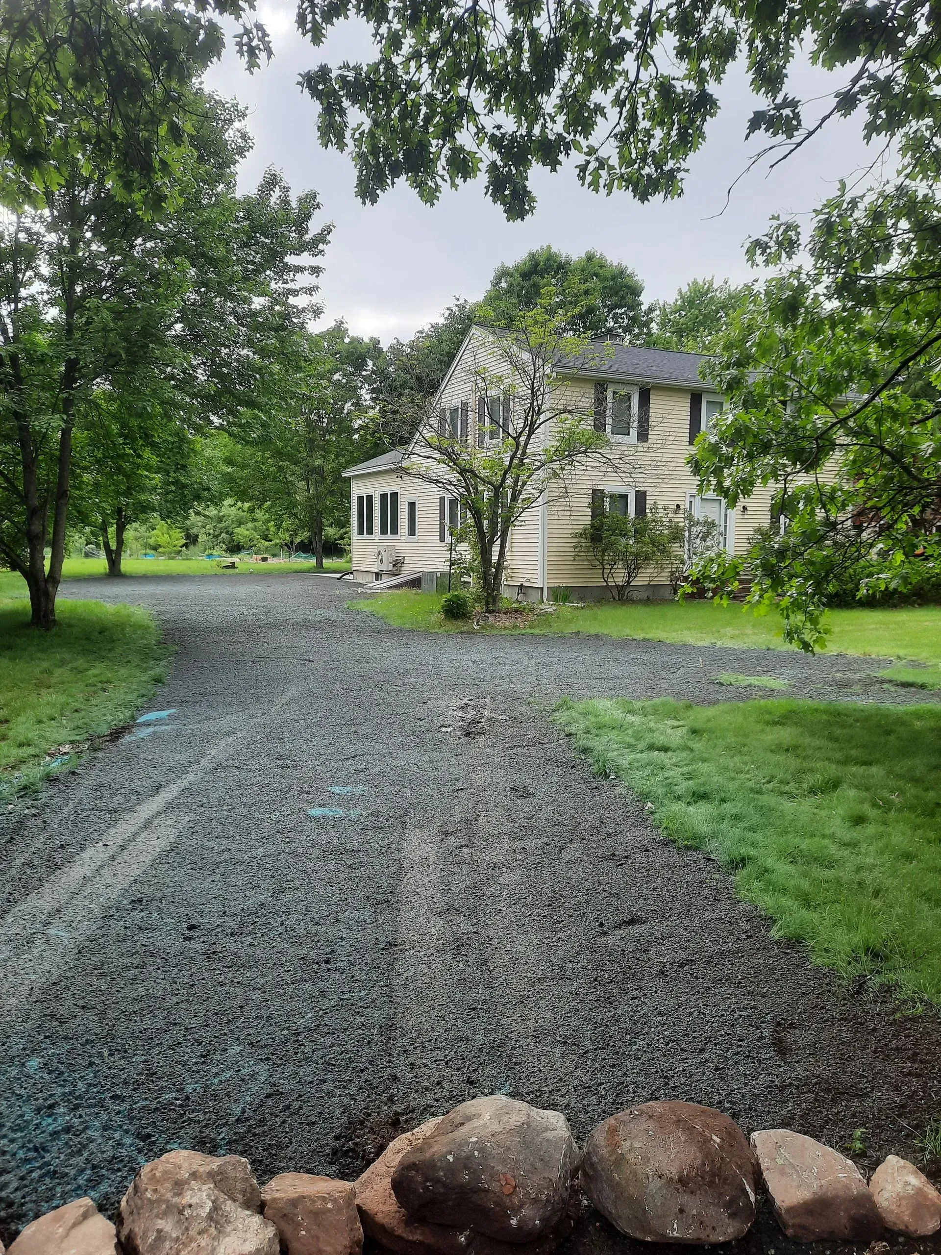 Gravel driveway leading to a white house amid green trees and lawn, viewed from a rock border in the foreground