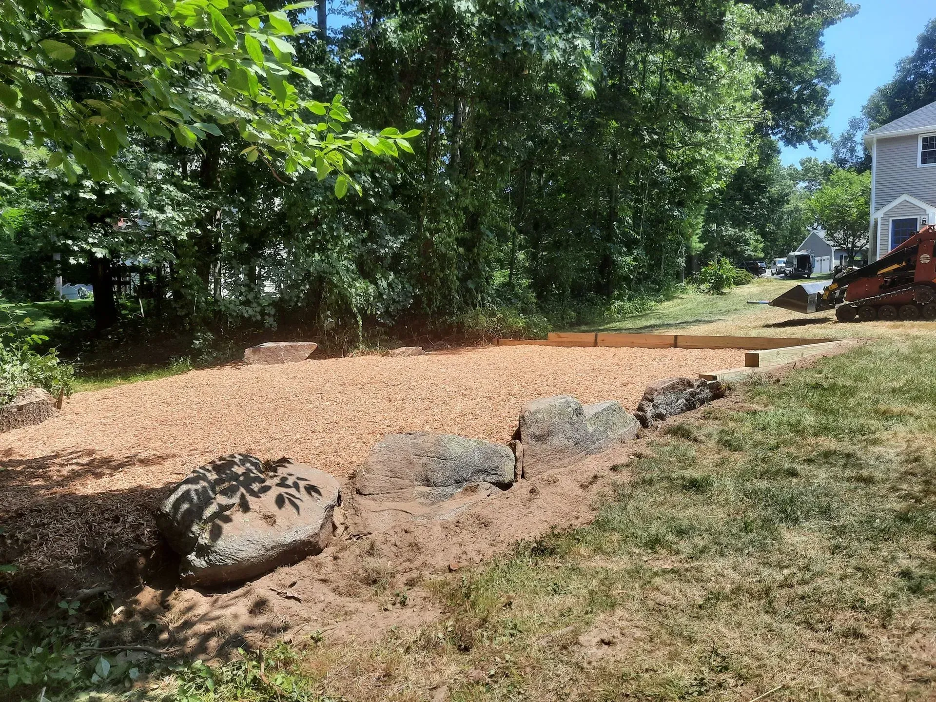 Wooded backyard with a gravel path, large rocks, and a house at the right edge