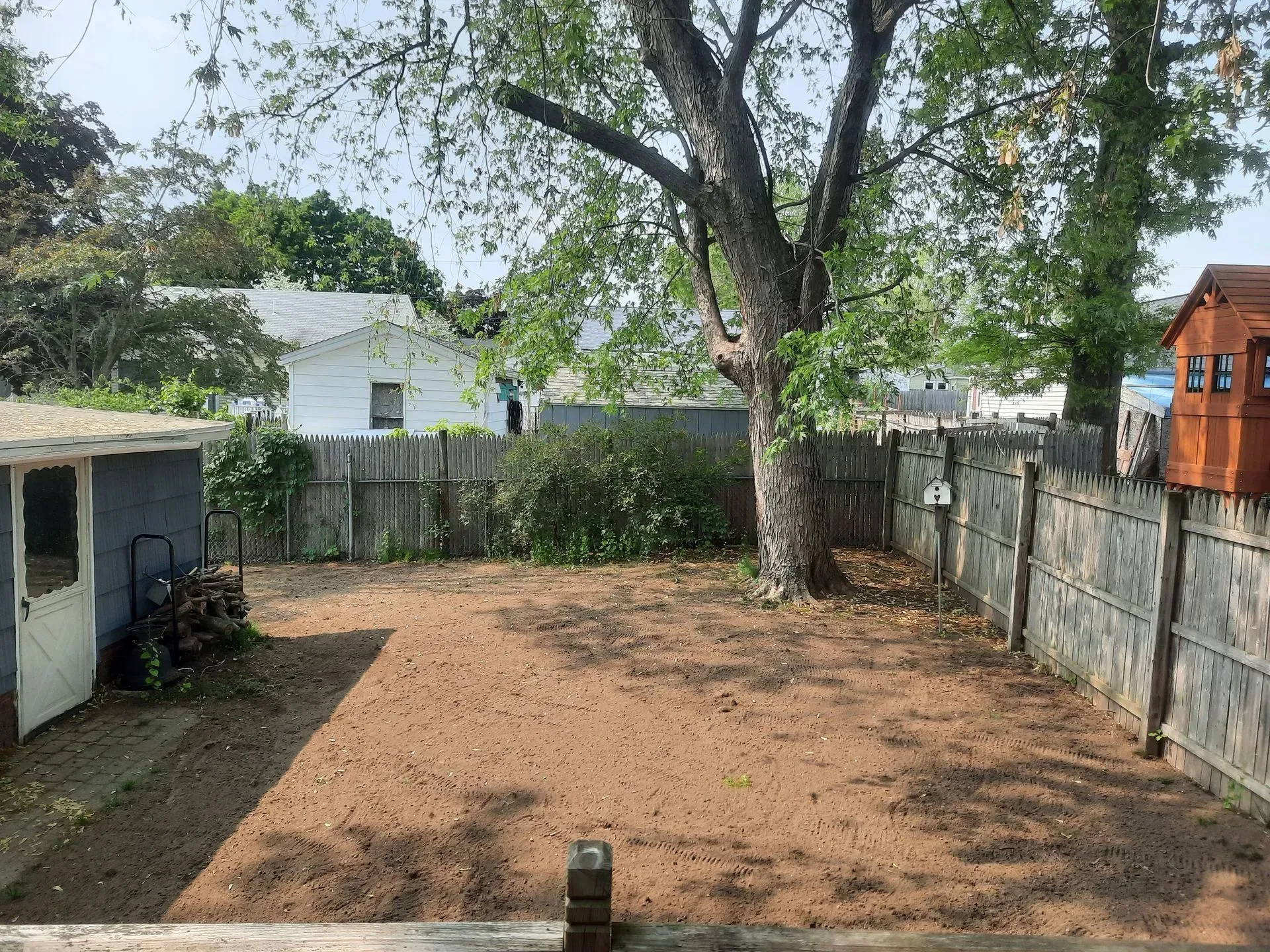 Empty dirt yard with a large tree, wooden fence, and small shed under a clear sky