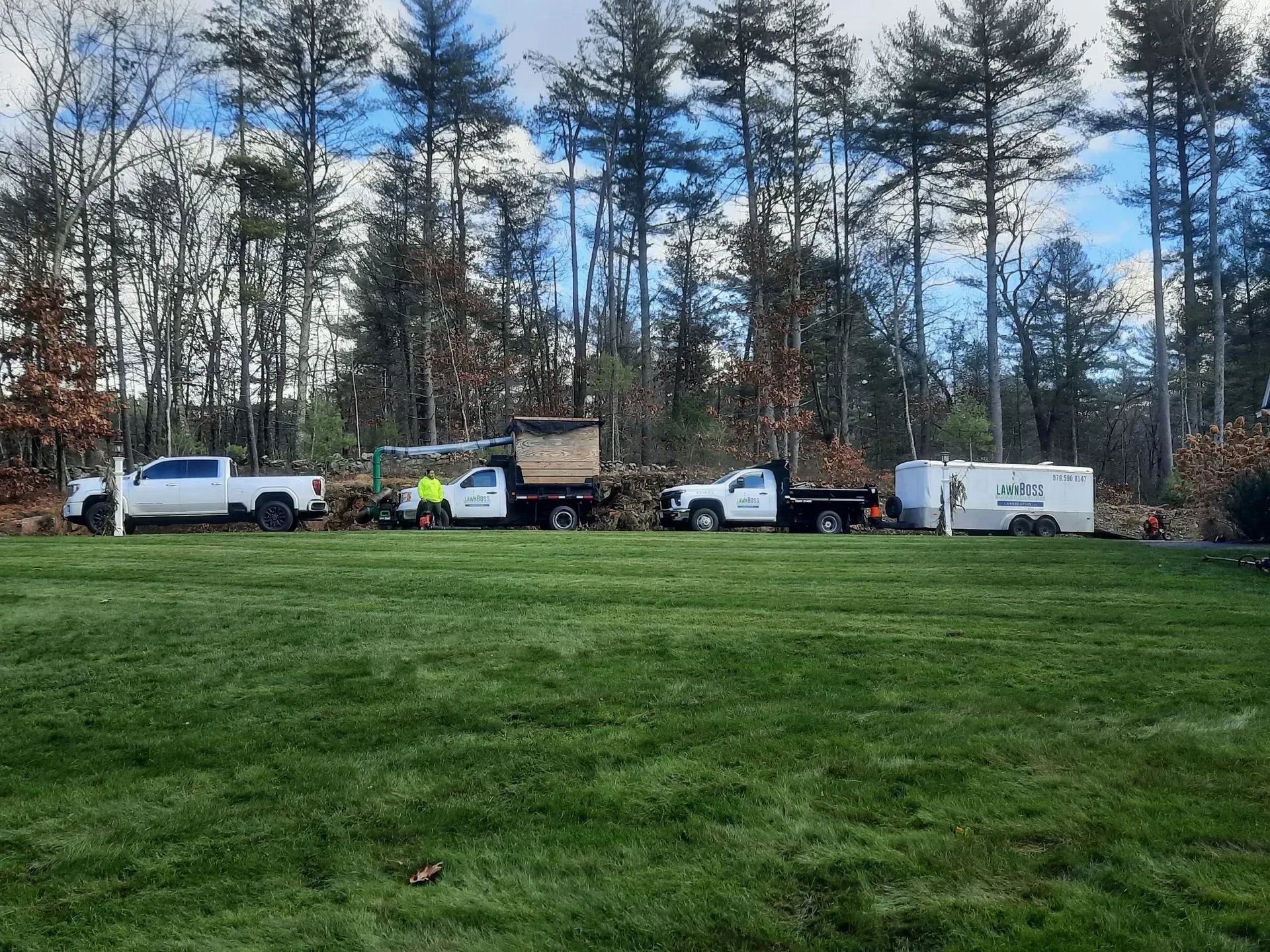 Utility vehicles lined up on a grassy field beside a wooded area under a cloudy sky.
