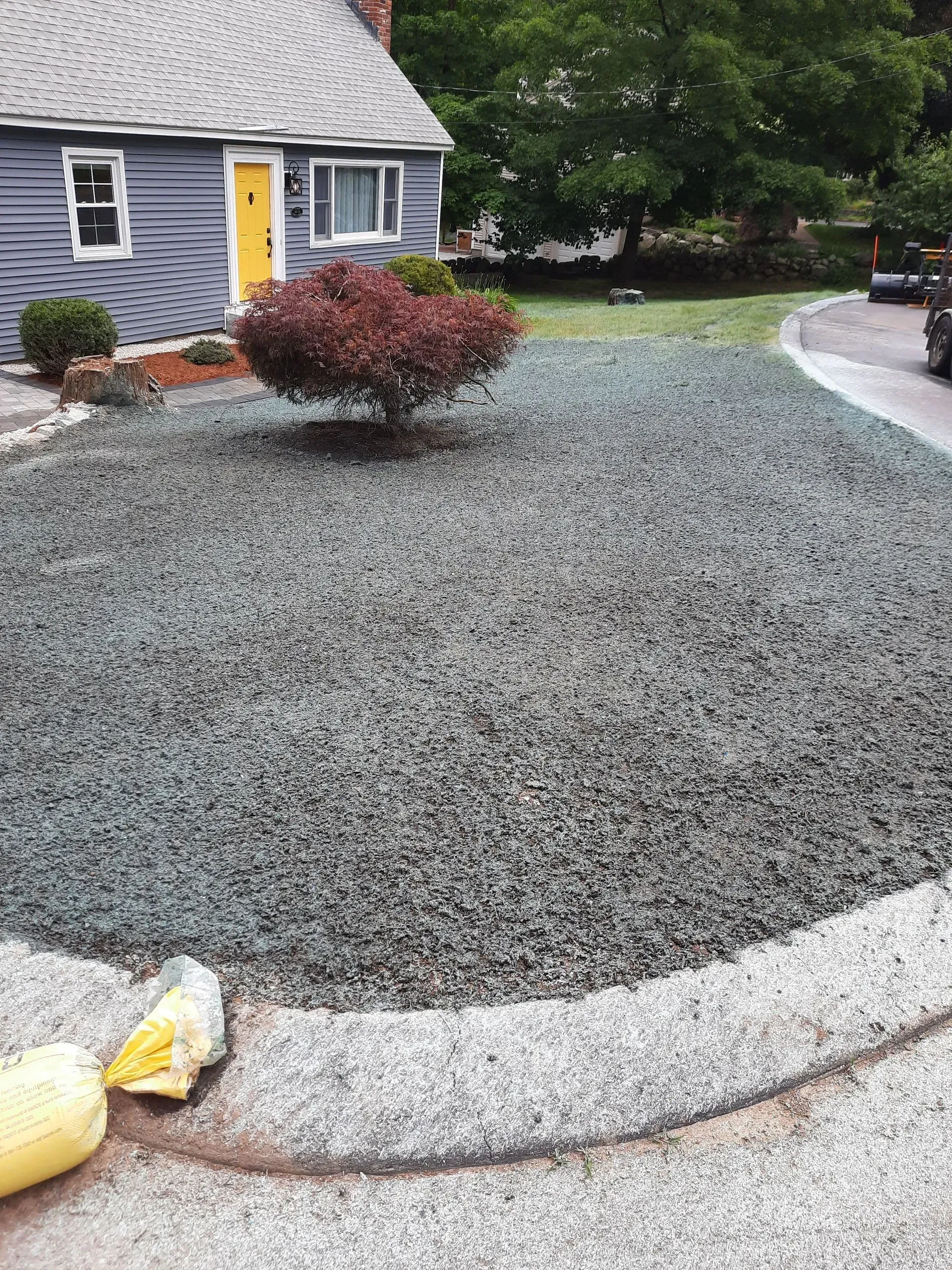 Gravel-covered front yard beside a gray house with a yellow door and shrubs, bordered by a curb.