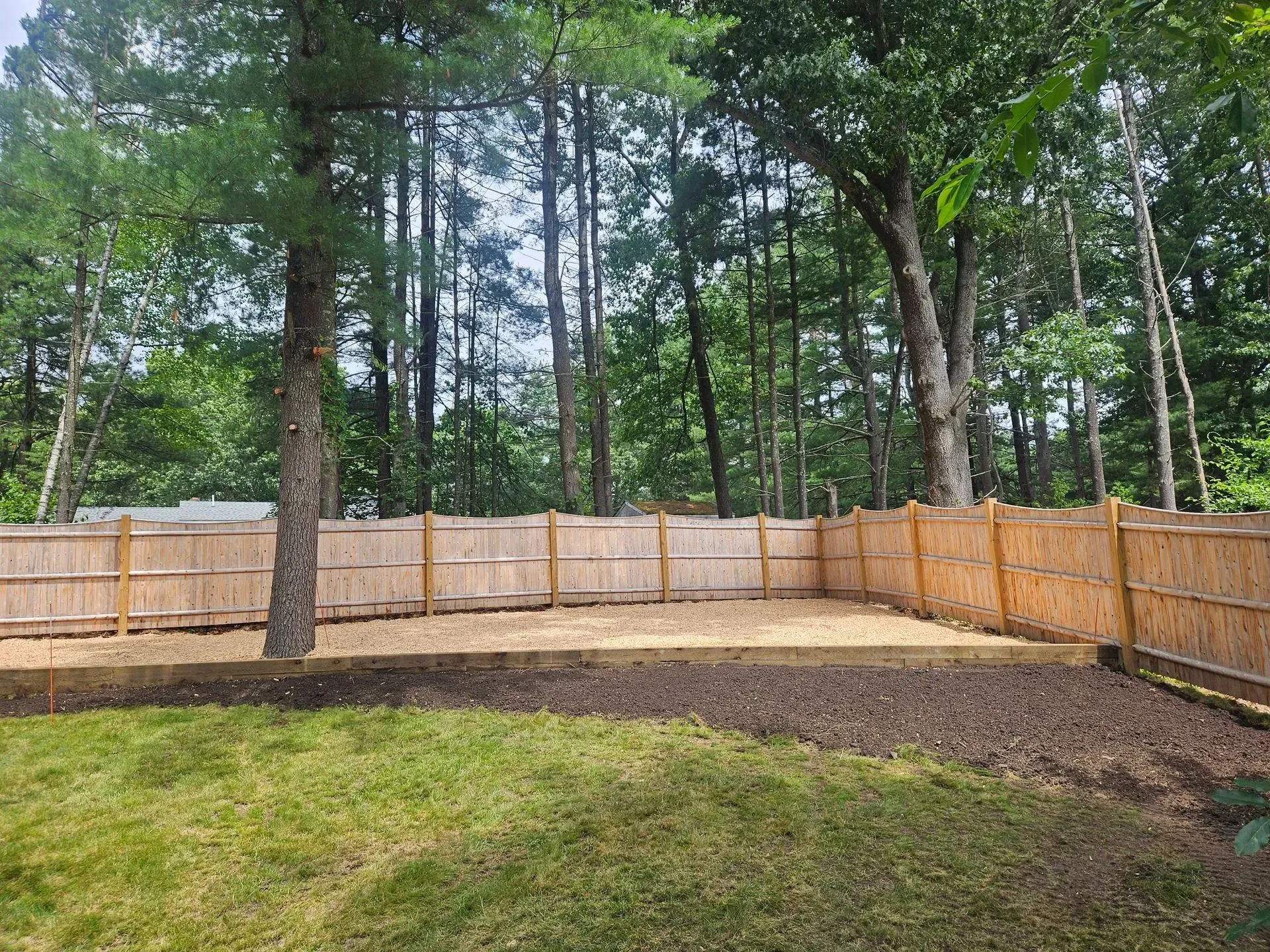 Wooden fenced backyard with fresh soil beds, grass, and tall trees beyond the fence.