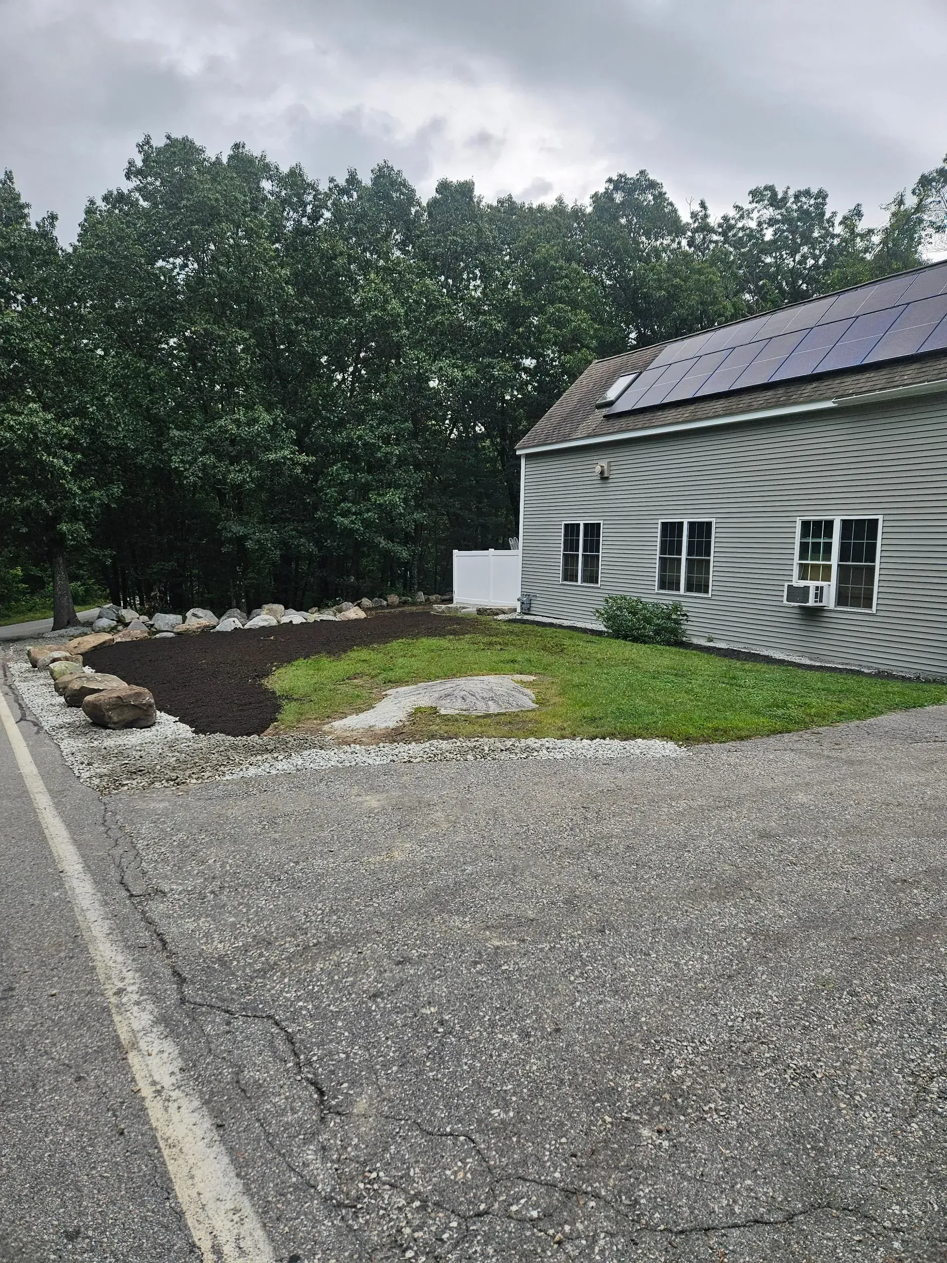 Gravel driveway beside a white house with solar panels, dark mulch landscaping, and trees in the background
