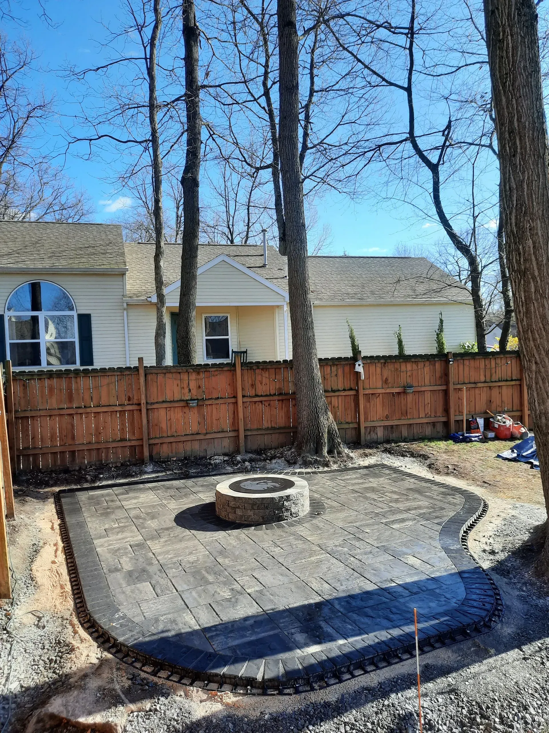Backyard patio with fire pit, wooden fence, and leafless trees under a blue sky