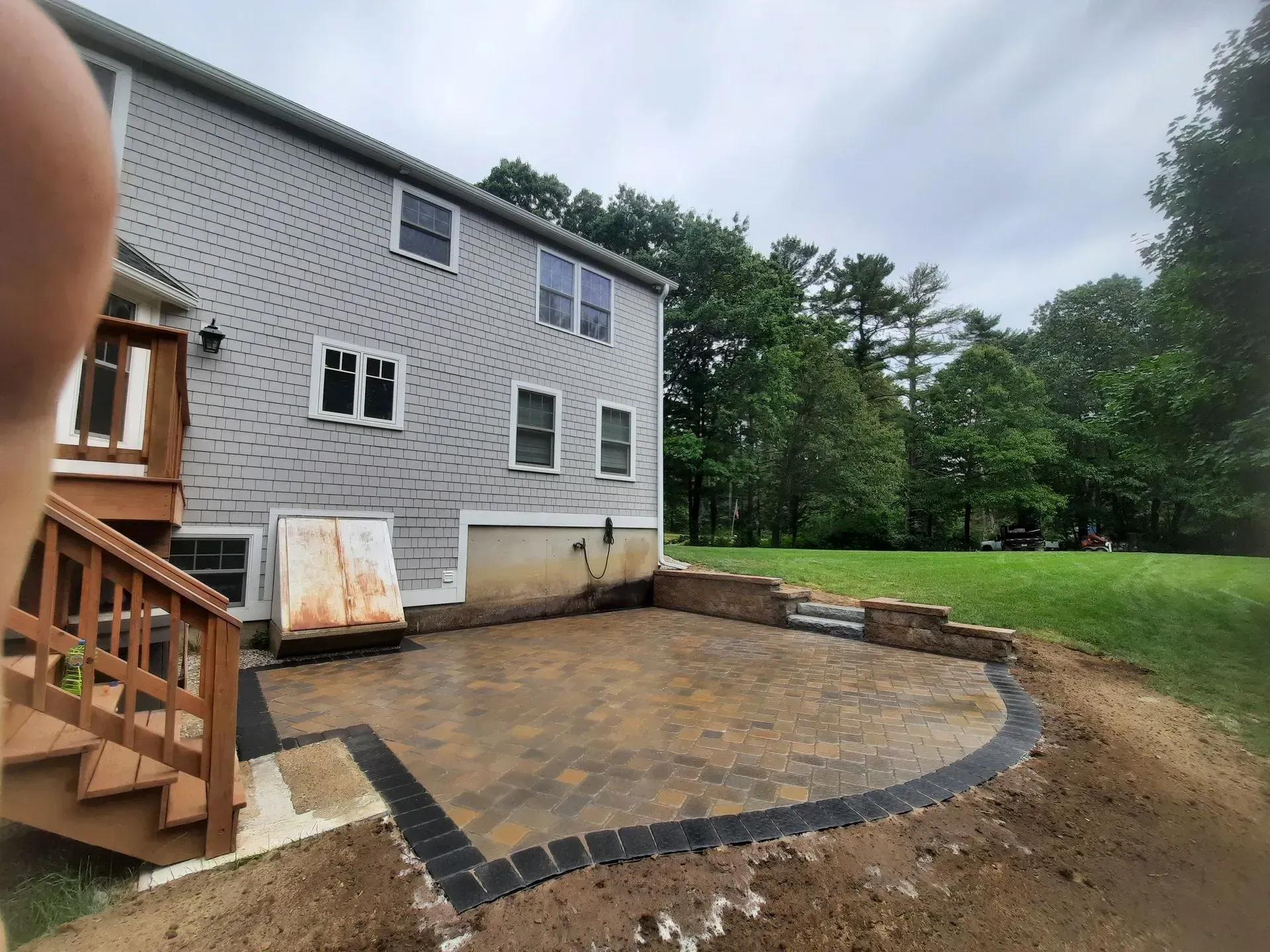 Backyard patio with pavers, curved stone border, and a gray house beside a lawn and trees