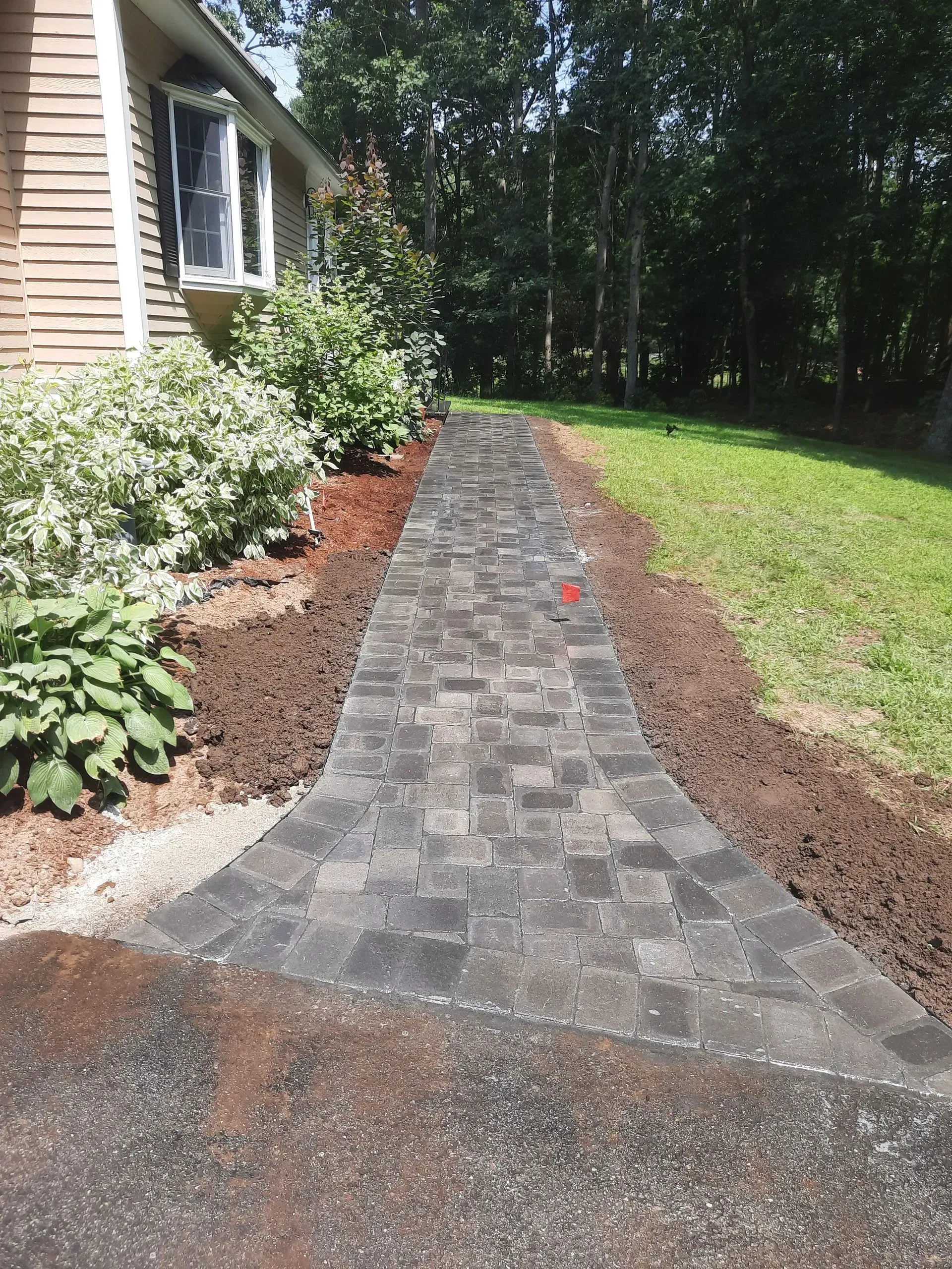 Brick garden walkway beside a house, bordered by shrubs and lawn under trees.