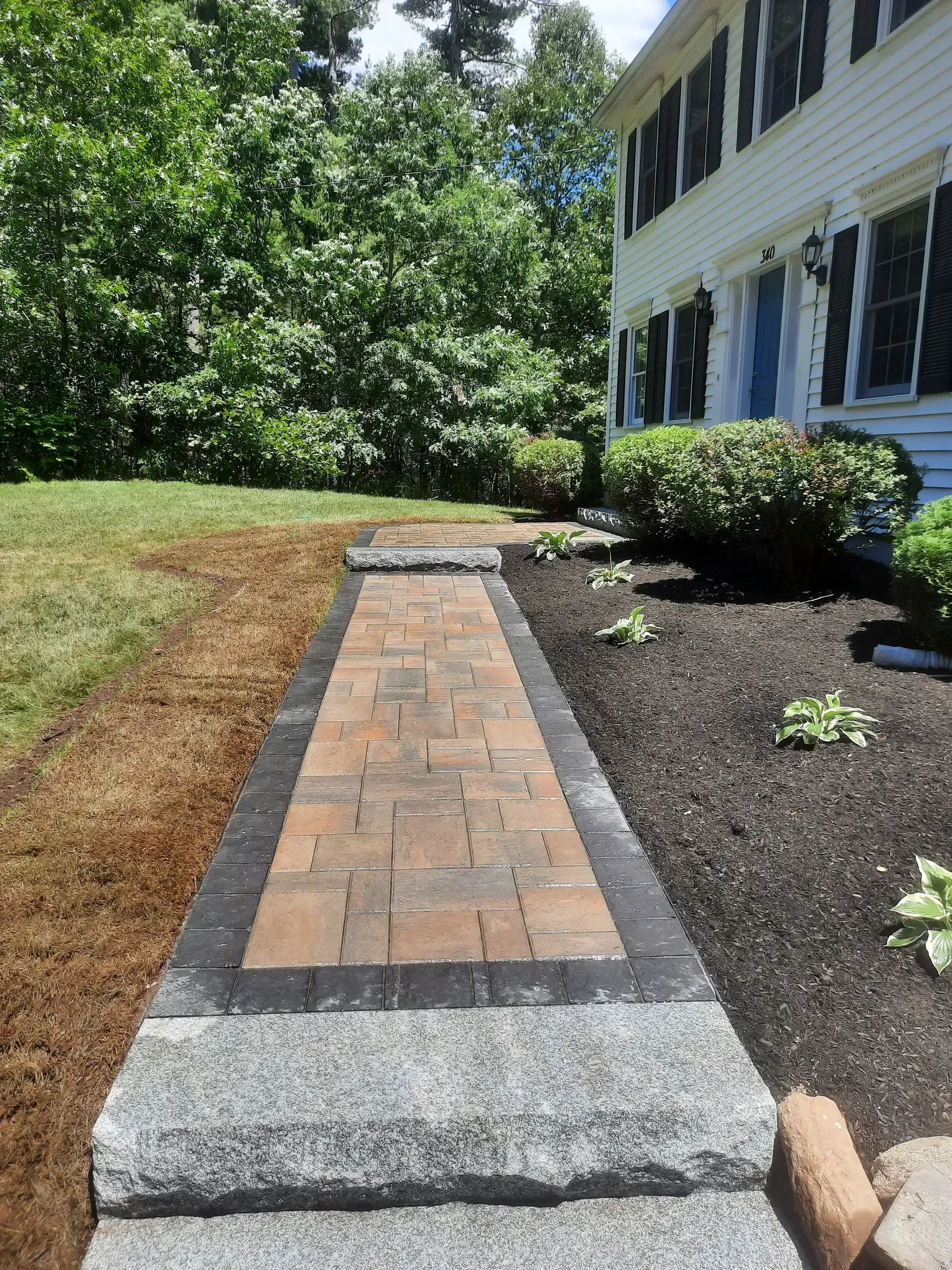 Brick walkway beside a white house, bordered by mulch and grass in a sunny yard.