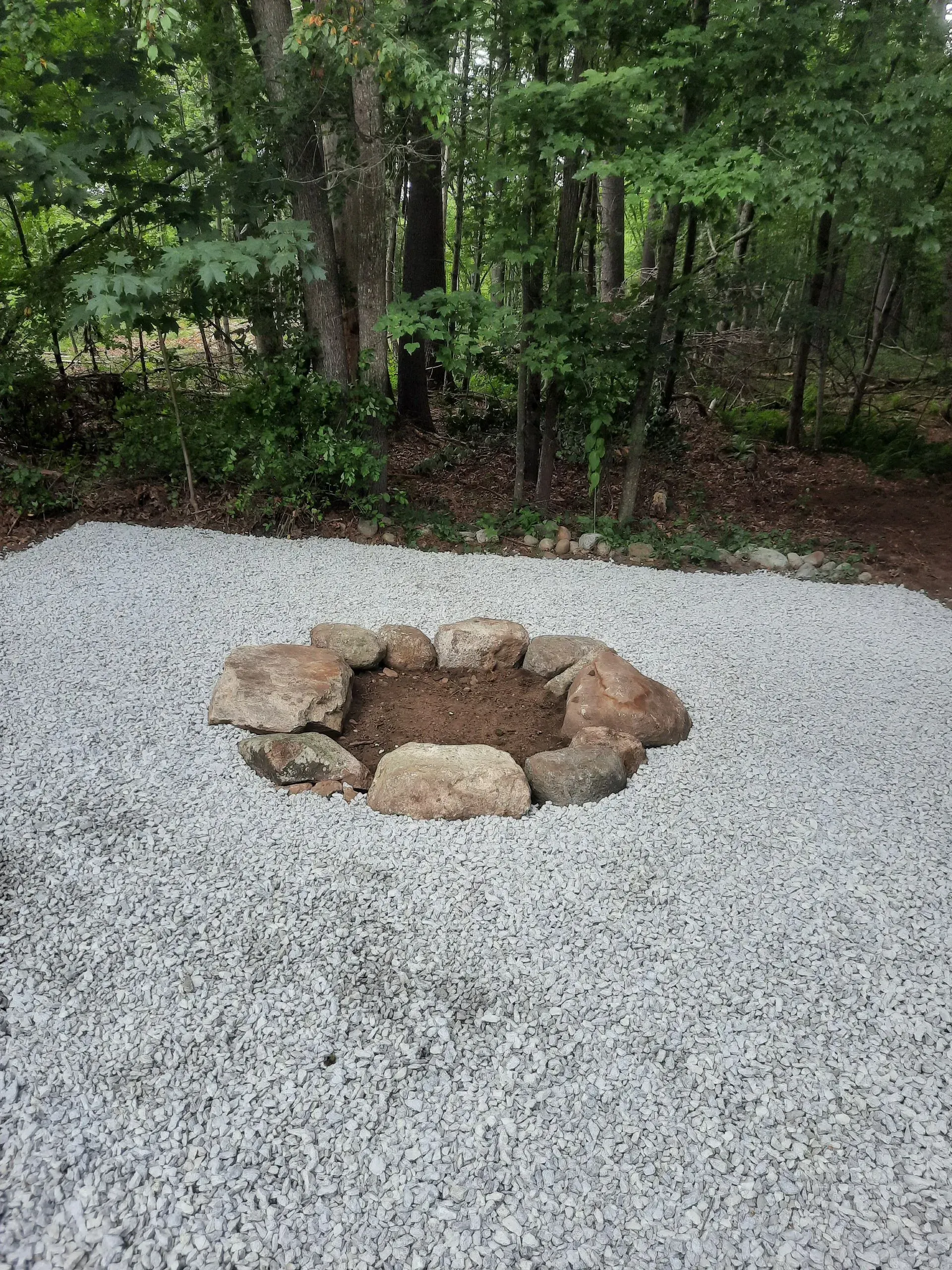 Stone fire pit on gravel beside a wooded area.