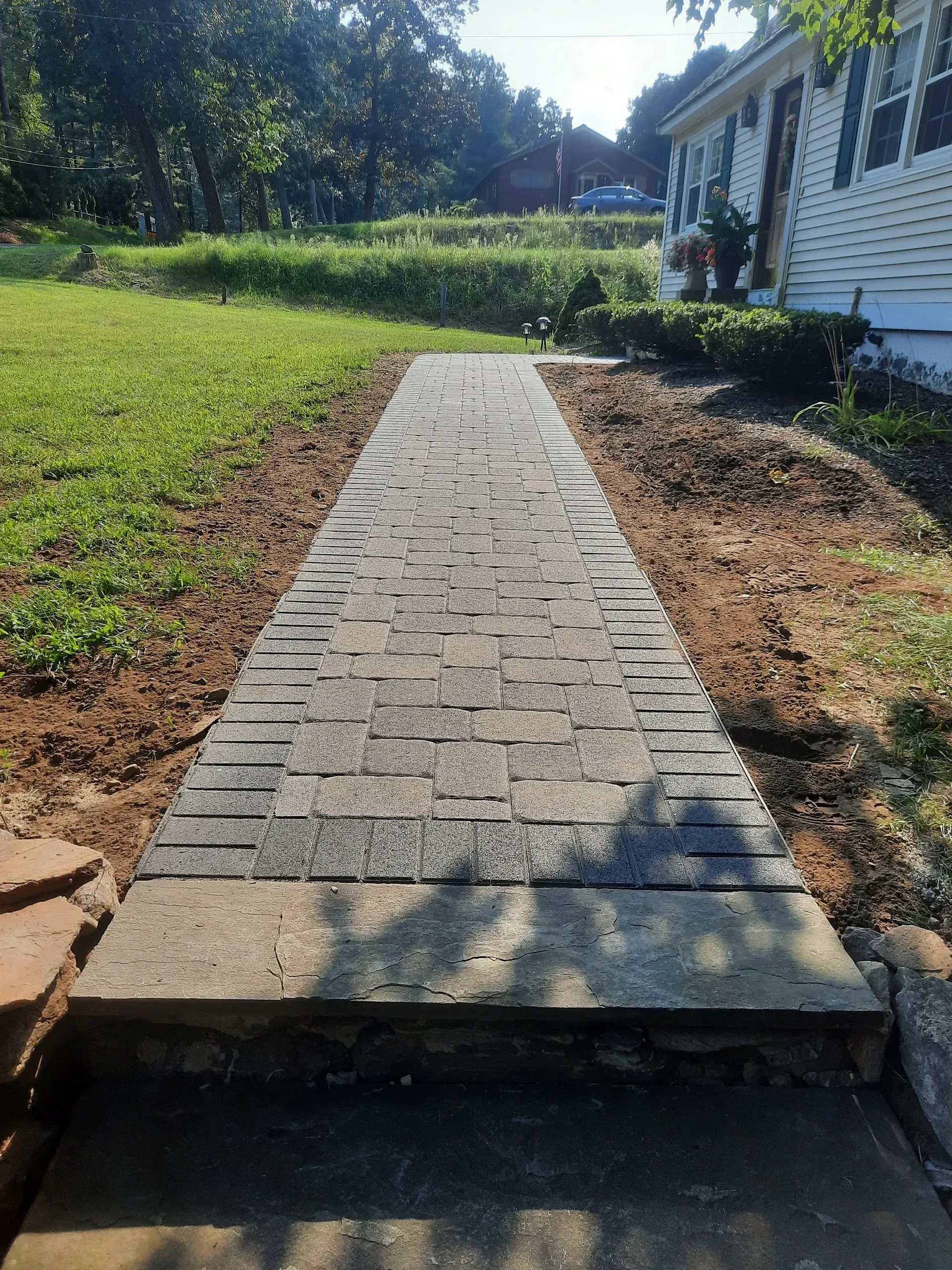 Brick walkway leading through a grassy yard beside a white house under bright sunlight.