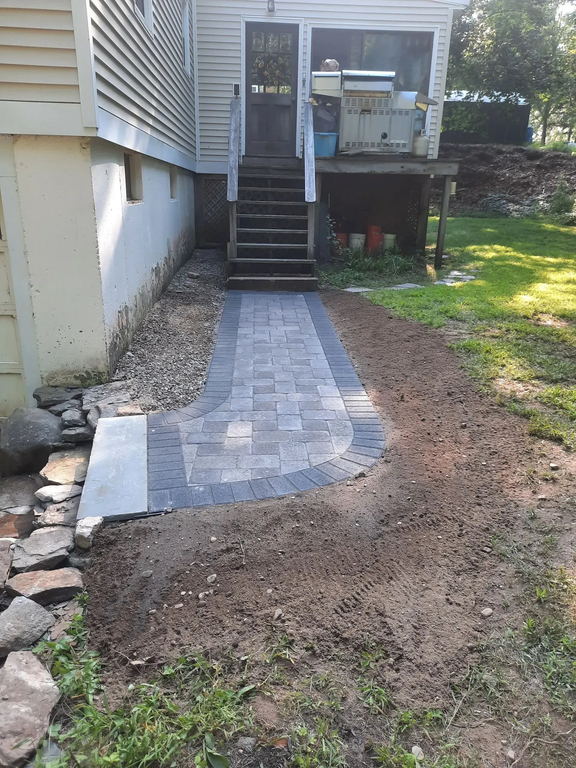 Freshly laid stone walkway leading to a house deck, bordered by gravel and grass, with stairs to the entry.