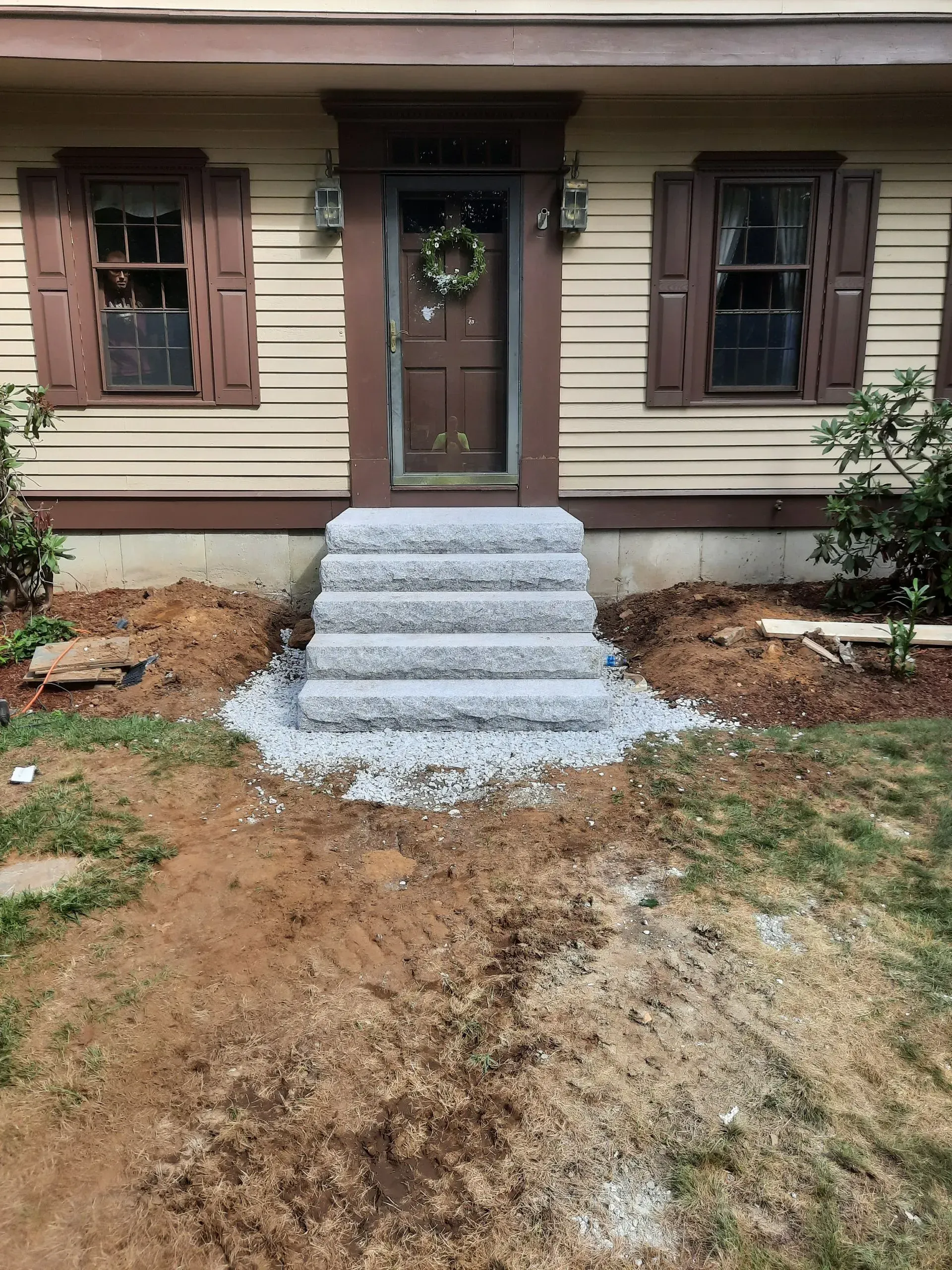Front entrance of a house with gray steps leading to a brown door and two windows, surrounded by dirt and gravel