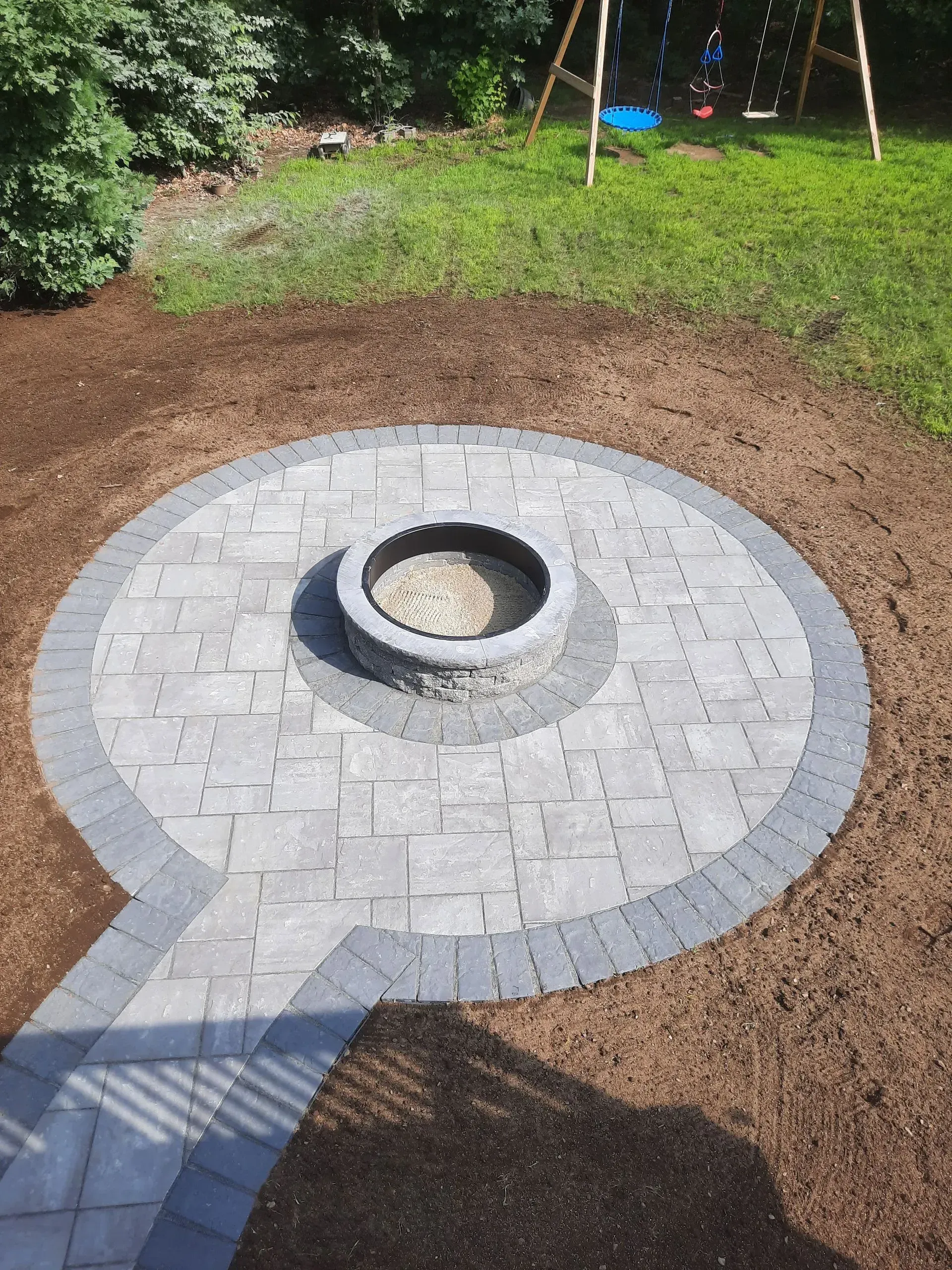 Circular stone fire pit set in a paved backyard patio beside a grassy lawn.