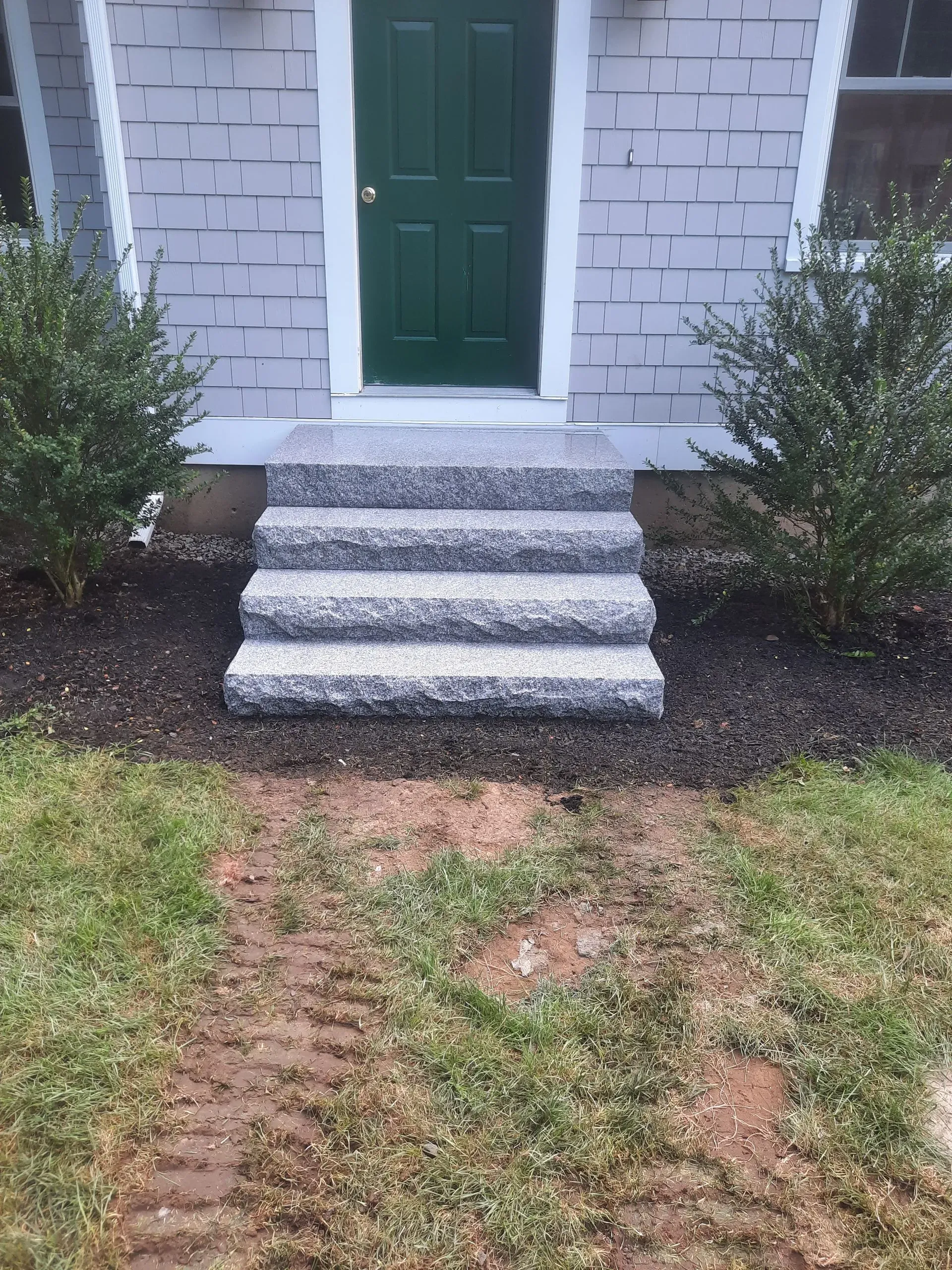 Front steps leading to a green door on a white brick house, with shrubs and a patchy lawn.