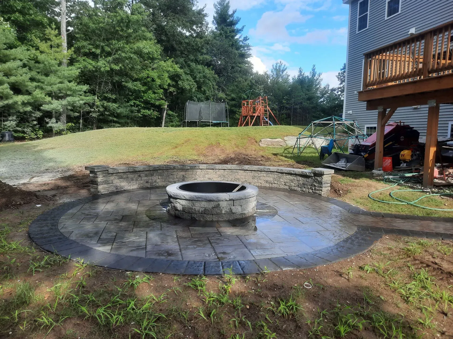 Backyard stone fire pit with curved paver patio near a wooden deck and grassy lawn