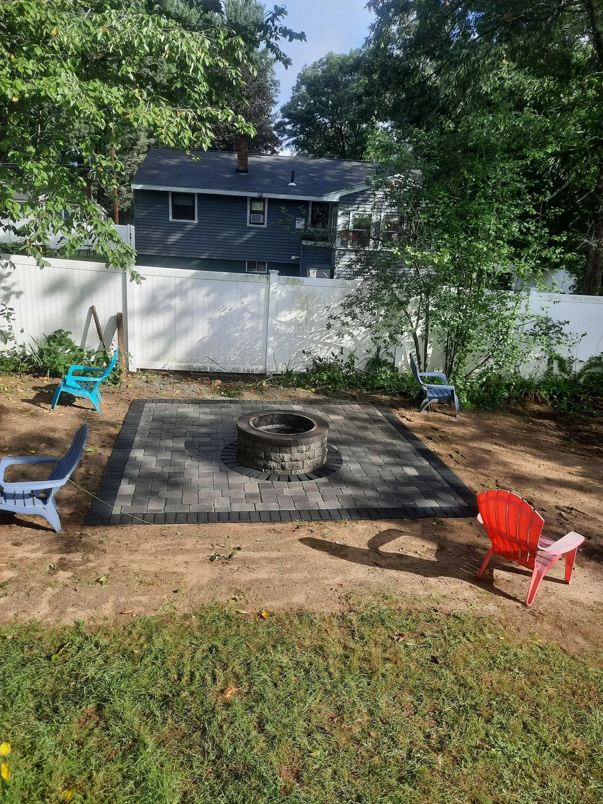 Backyard fire pit with paver patio, surrounded by trees, white fence, and two small chairs
