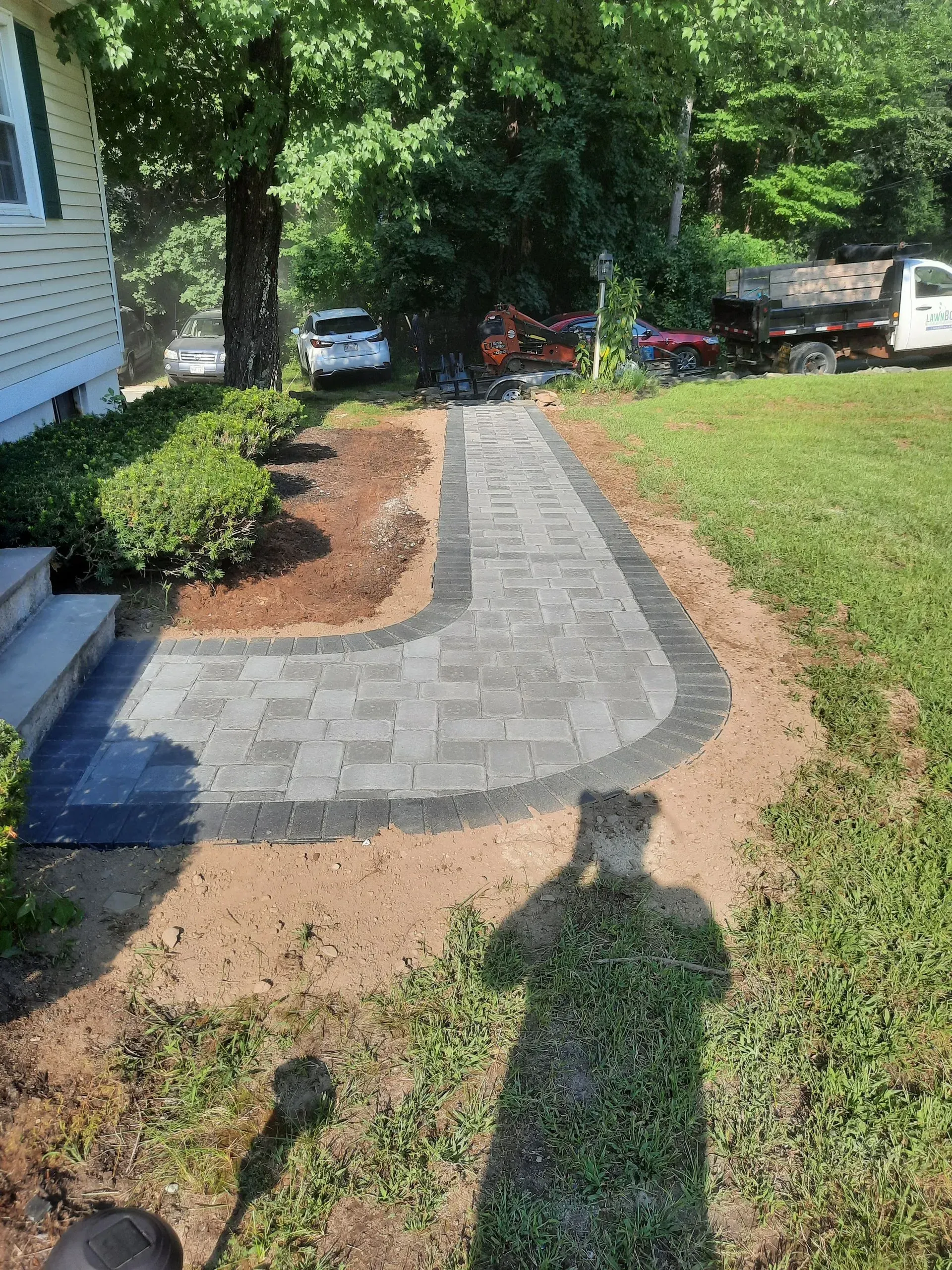 New gray paver walkway beside a house, curving through a landscaped yard with trees and mulch.