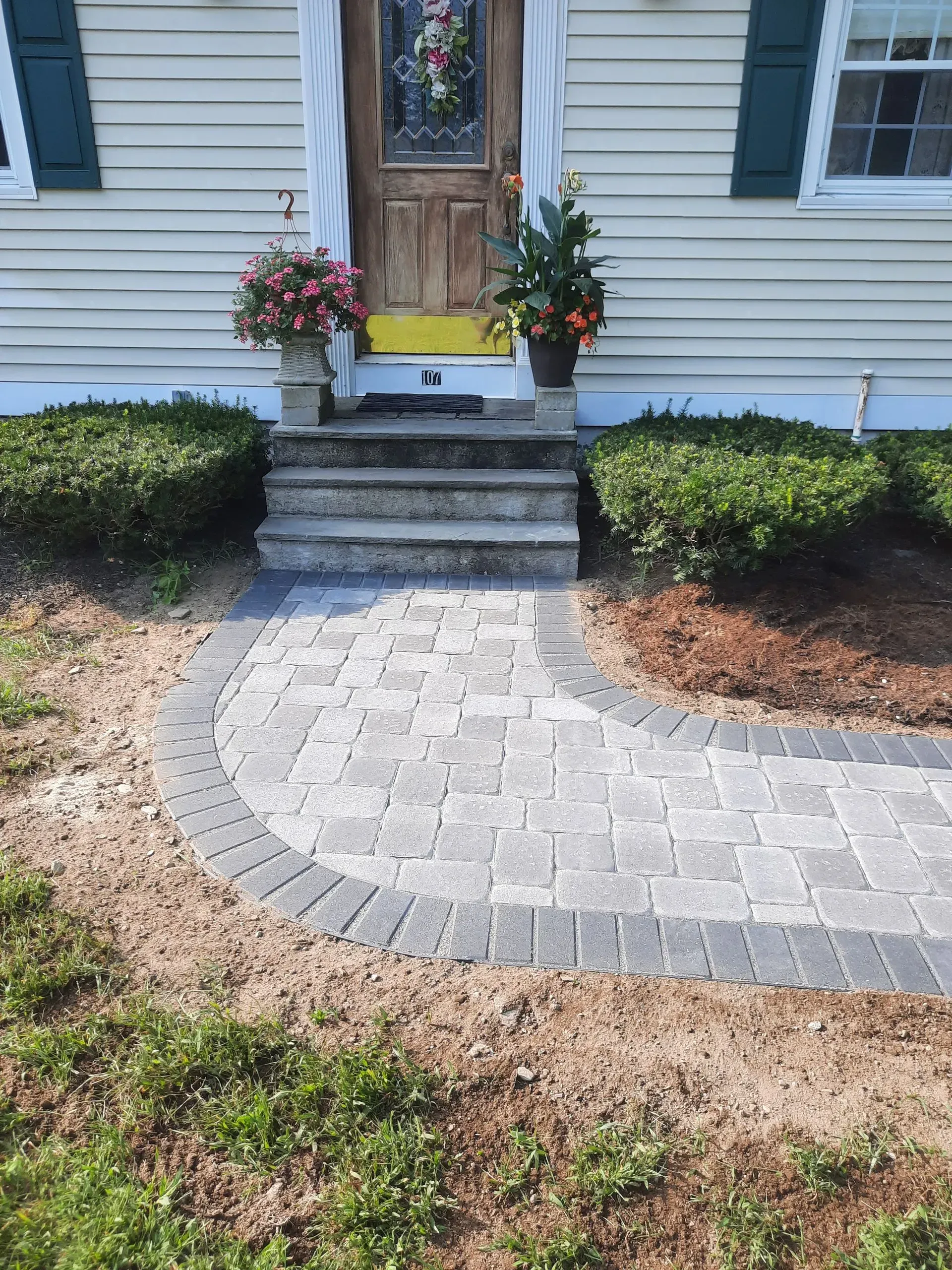 Front walkway of gray pavers leading to house steps and a wooden front door with flower pots and shrubs nearby