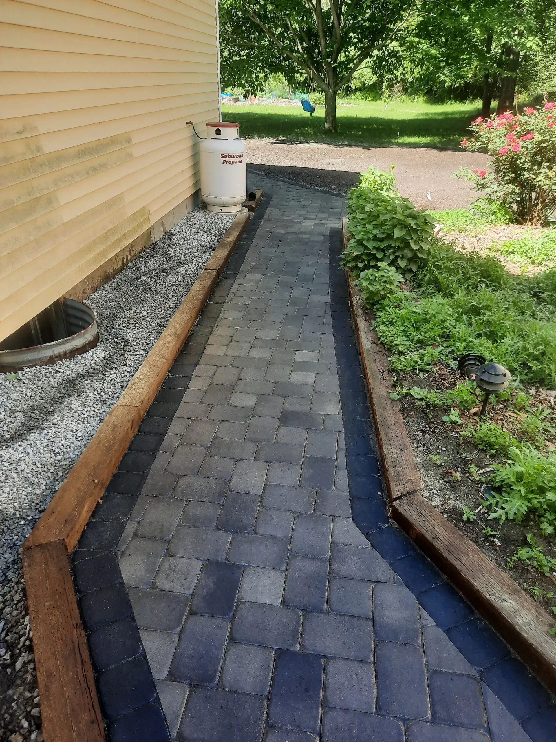 Narrow brick walkway beside a house, with a gravel border and green garden plants.
