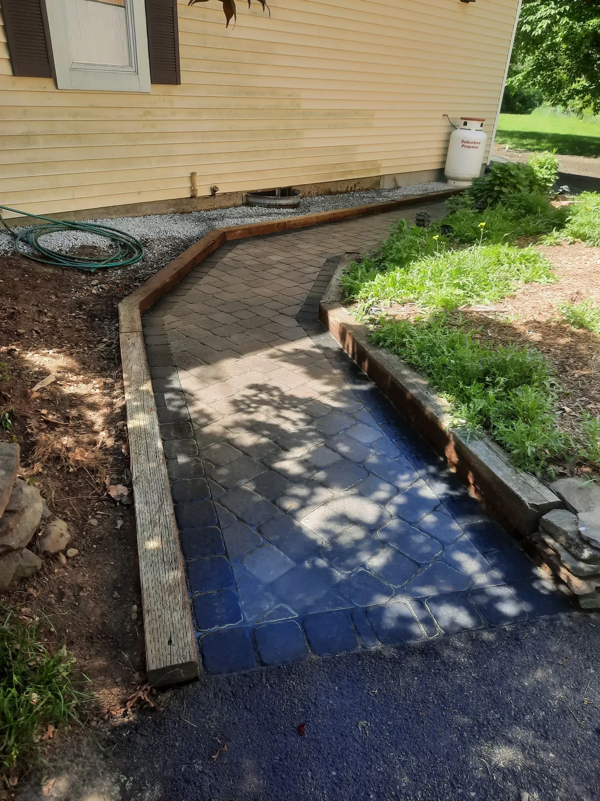 Curved brick-bordered walkway beside a house, with a blue-tiled path and green garden beds.