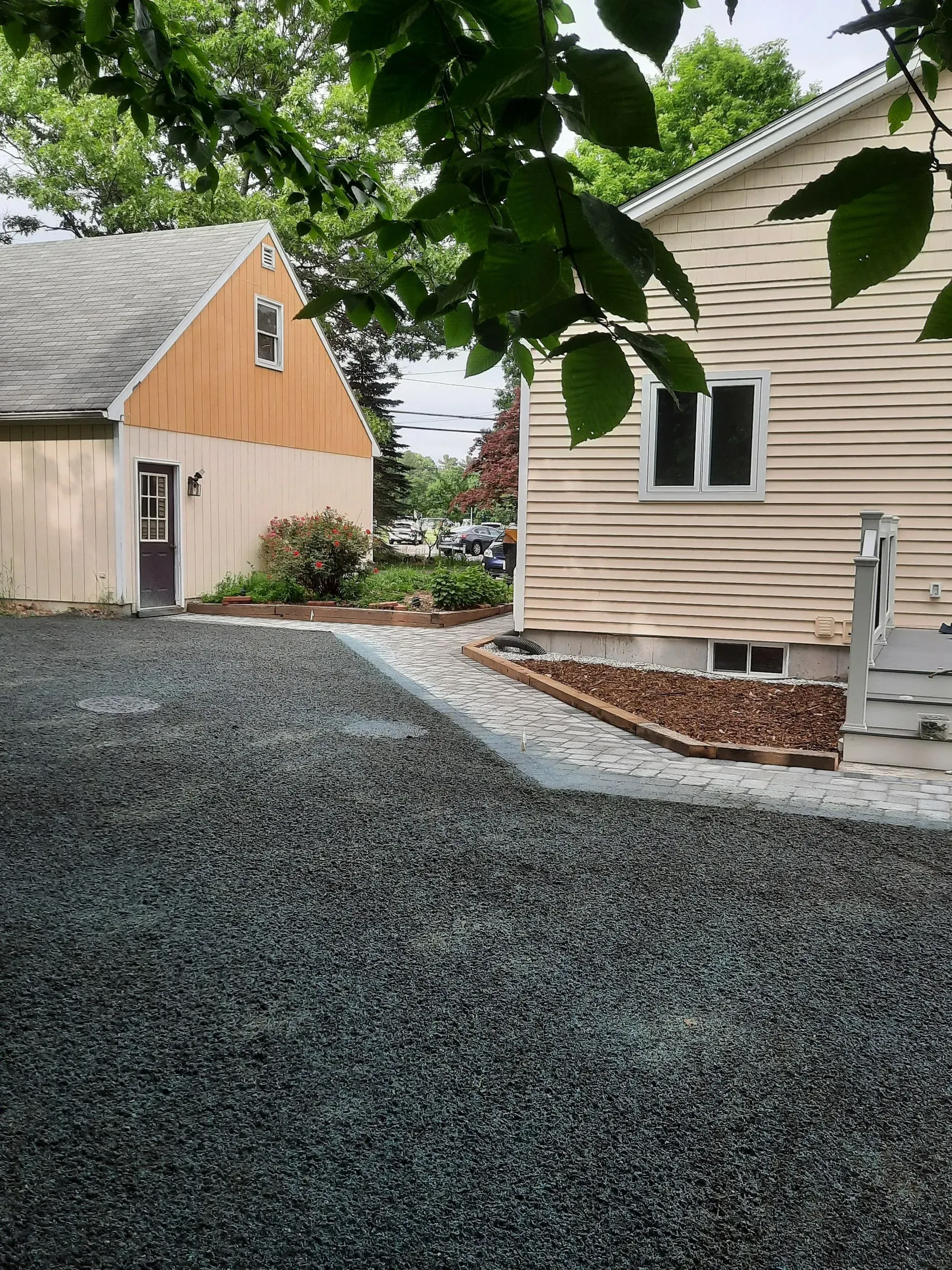 Gravel driveway between two beige buildings, with trees overhead and white patio chair on the right.