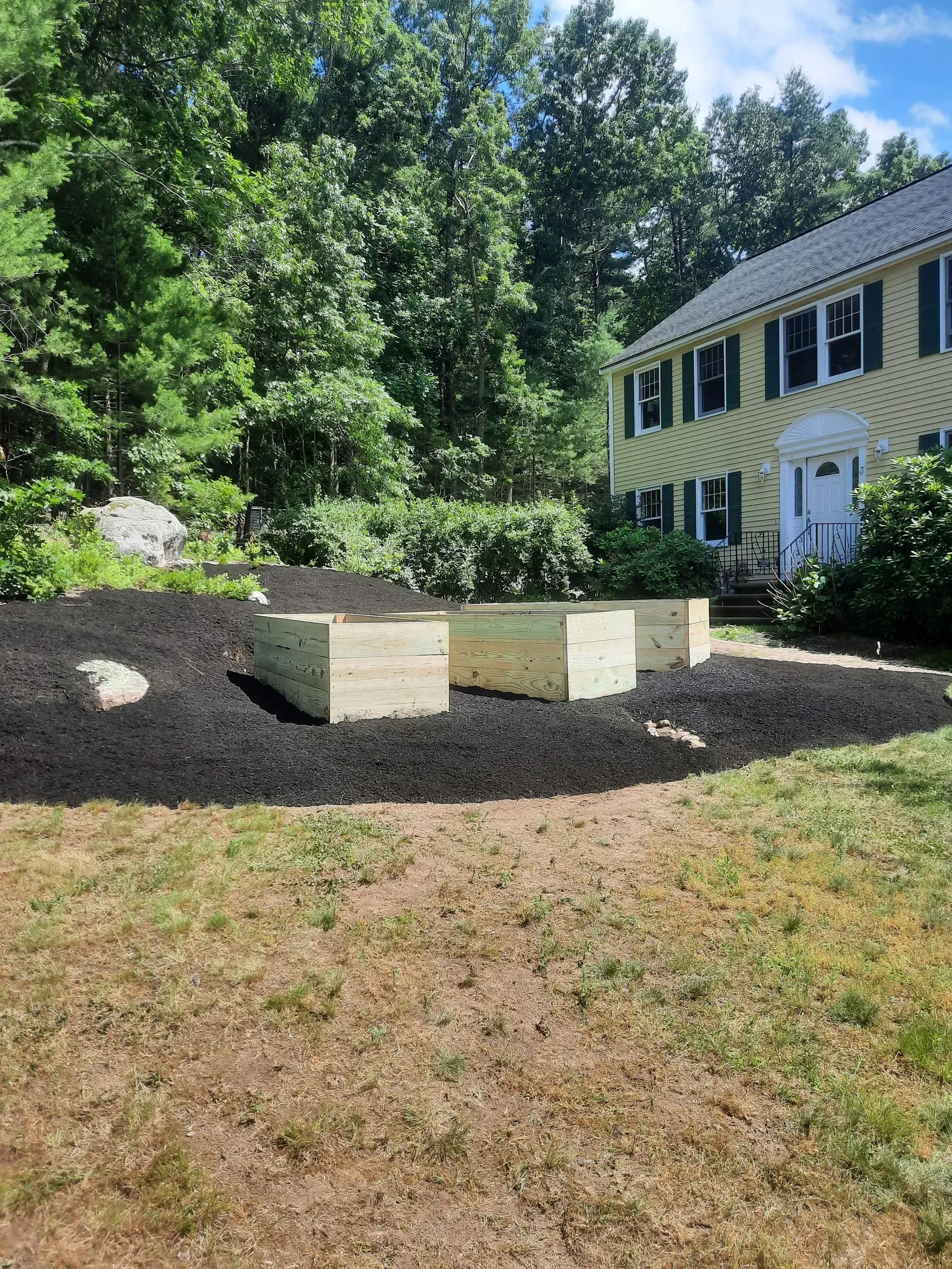 Front yard landscaping with dark mulch, three stone planters, and a yellow house beside trees