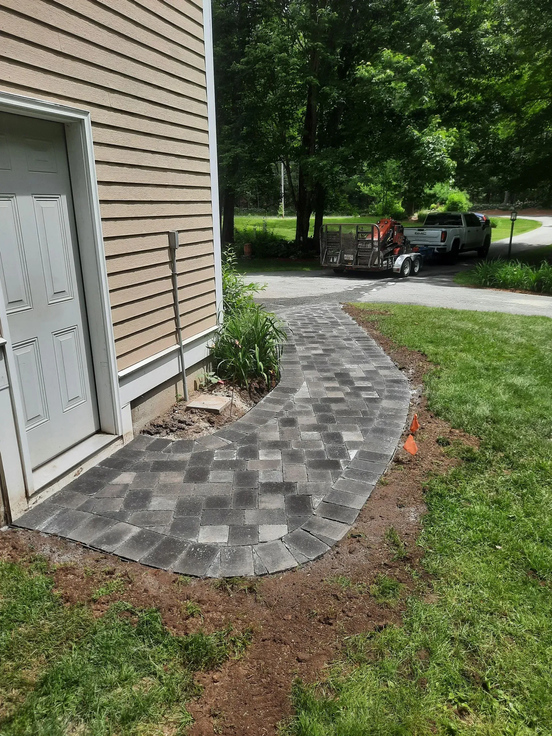 Curved gray paver walkway beside a house, leading to a driveway with a parked car and trees beyond.
