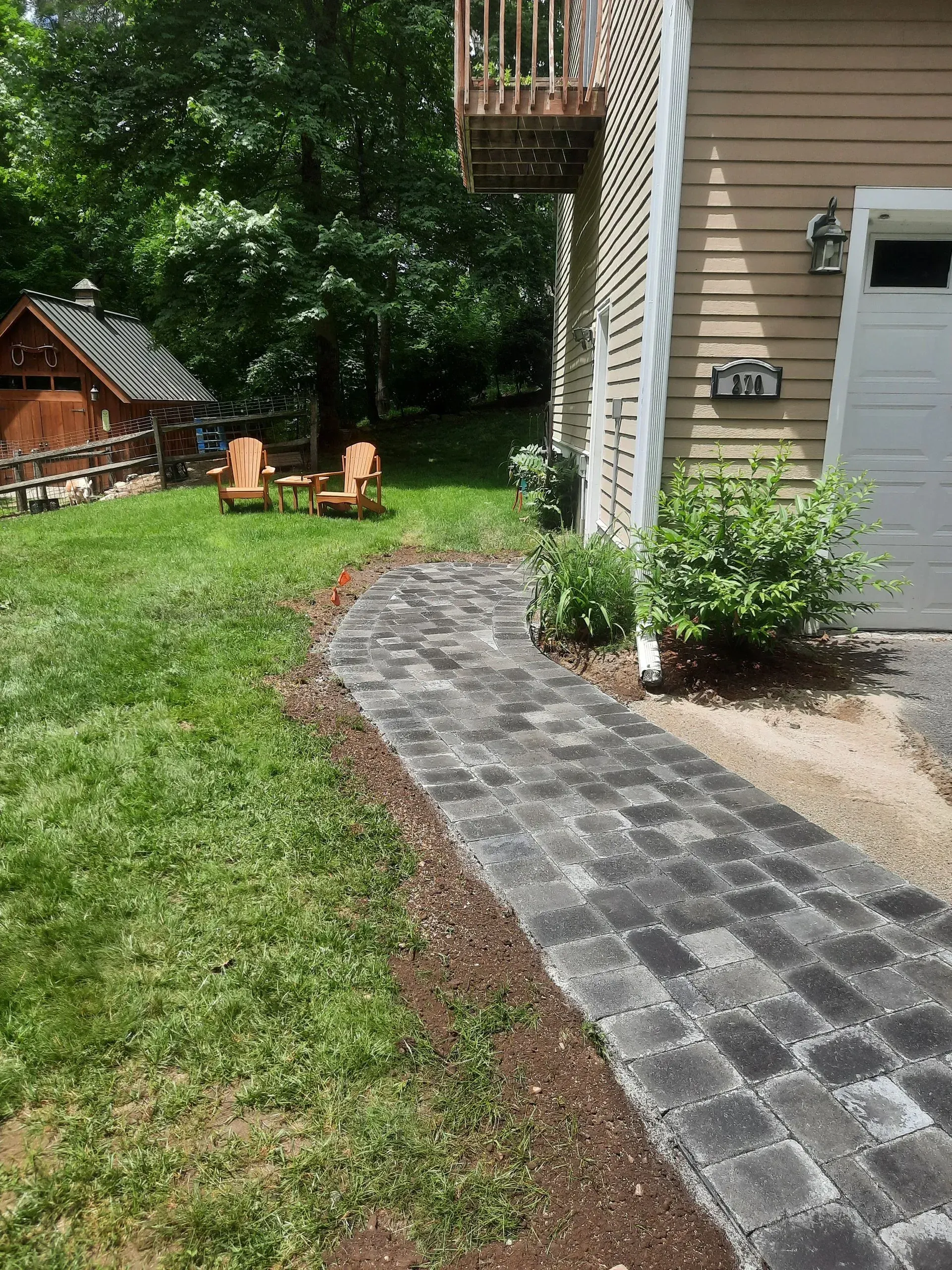 Curved stone walkway beside a house, leading to two orange chairs in a green backyard.