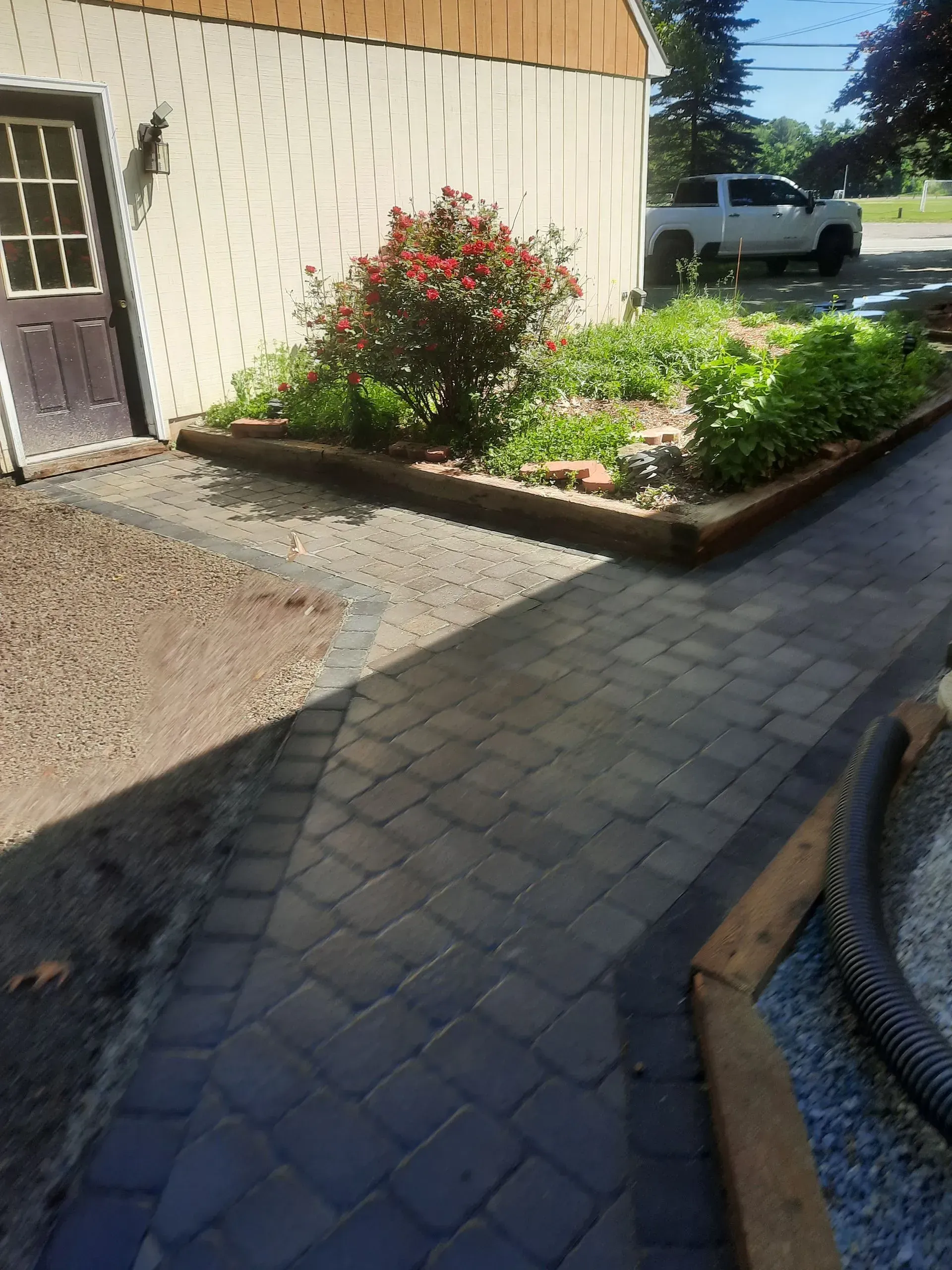 Brick walkway beside a house with a raised flower bed and gravel driveway, with a white truck in the background.