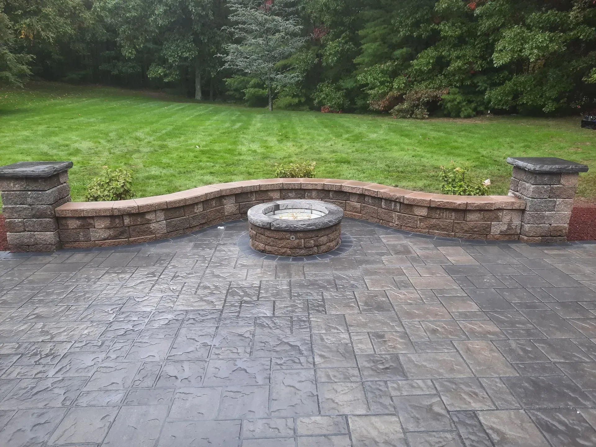 Stone patio with a curved brick fire pit wall and a green lawn and trees beyond.