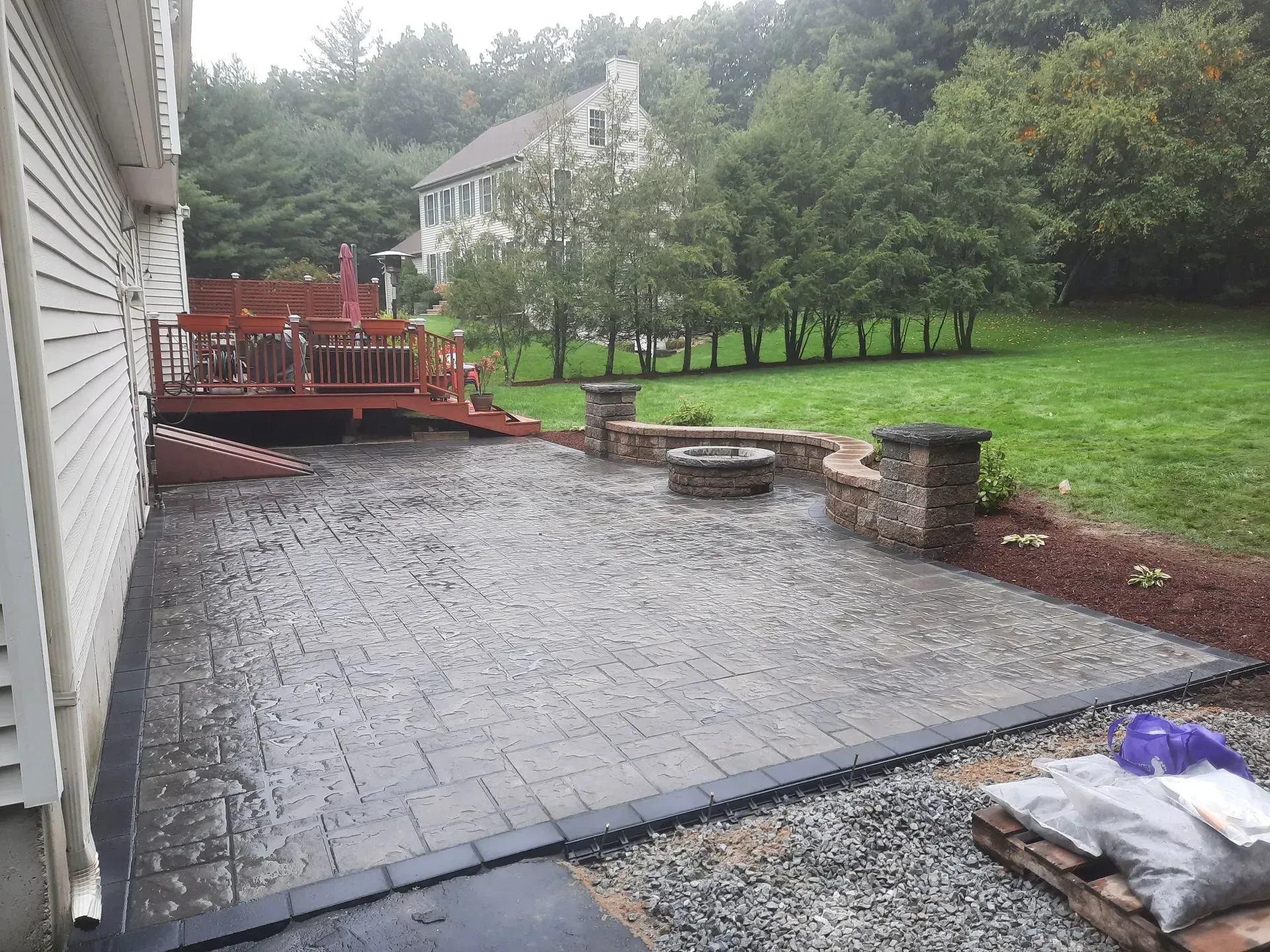 Rainy backyard patio with red deck, wet stone pavers, and a grassy yard beside a house
