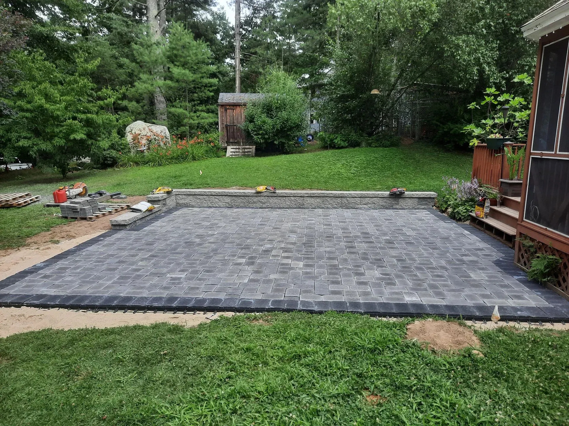 Backyard patio with gray pavers, bordered by grass and trees, beside a shed and garden area