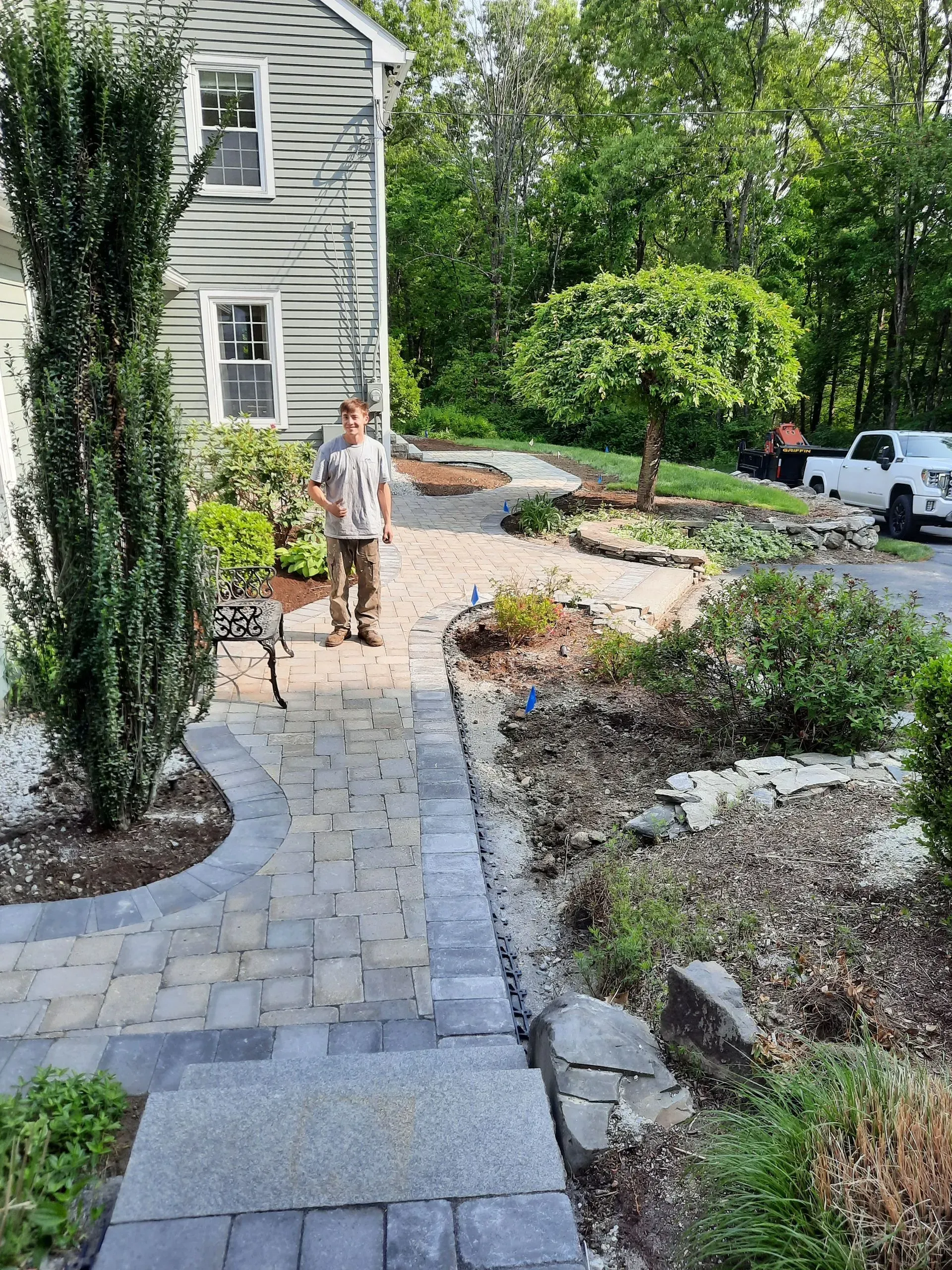 Stone walkway beside a house under construction, with a worker standing near landscaping stones and shrubs.