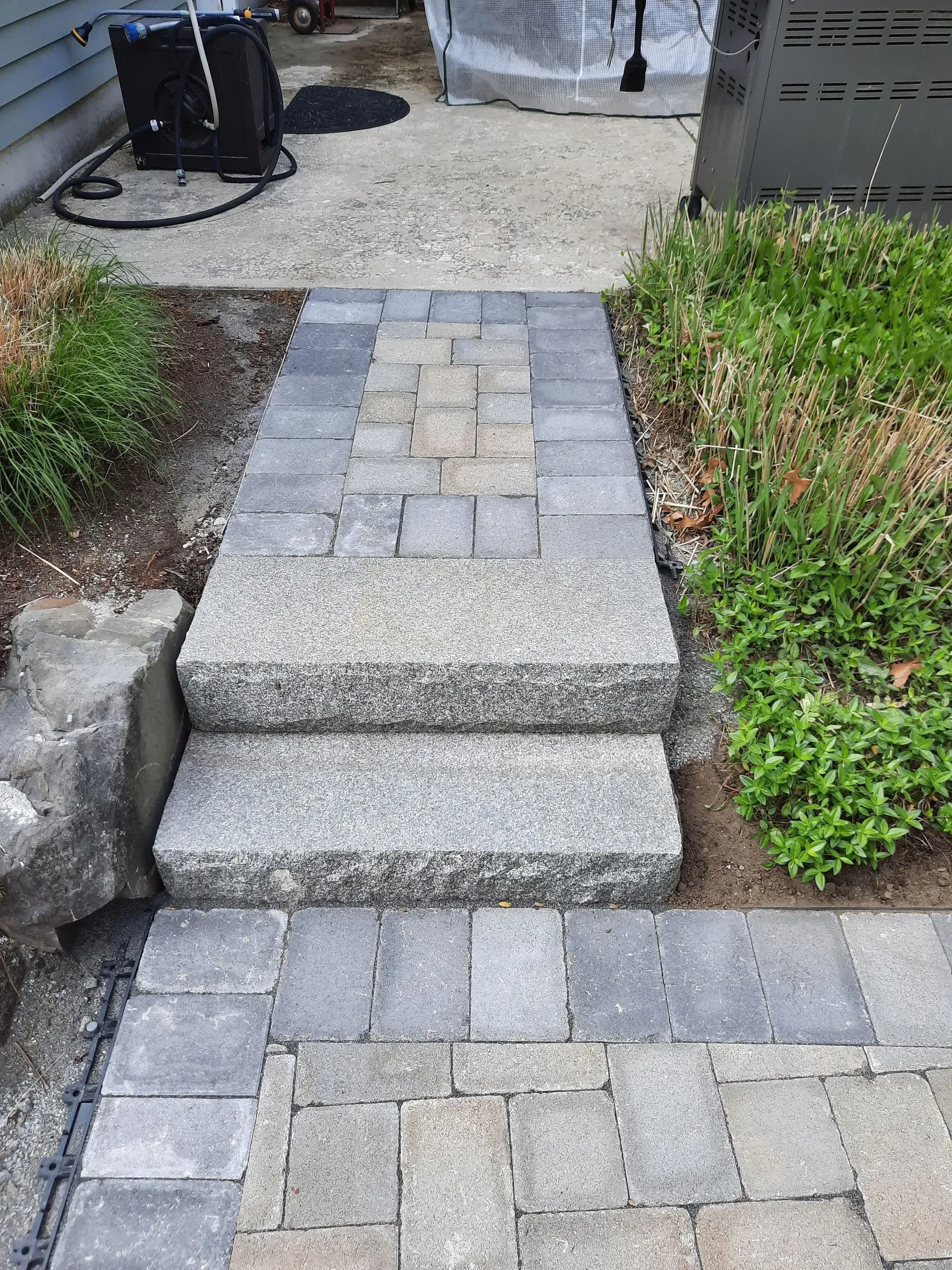 Stone steps leading from a paver walkway to a gravel patio, bordered by green shrubs.