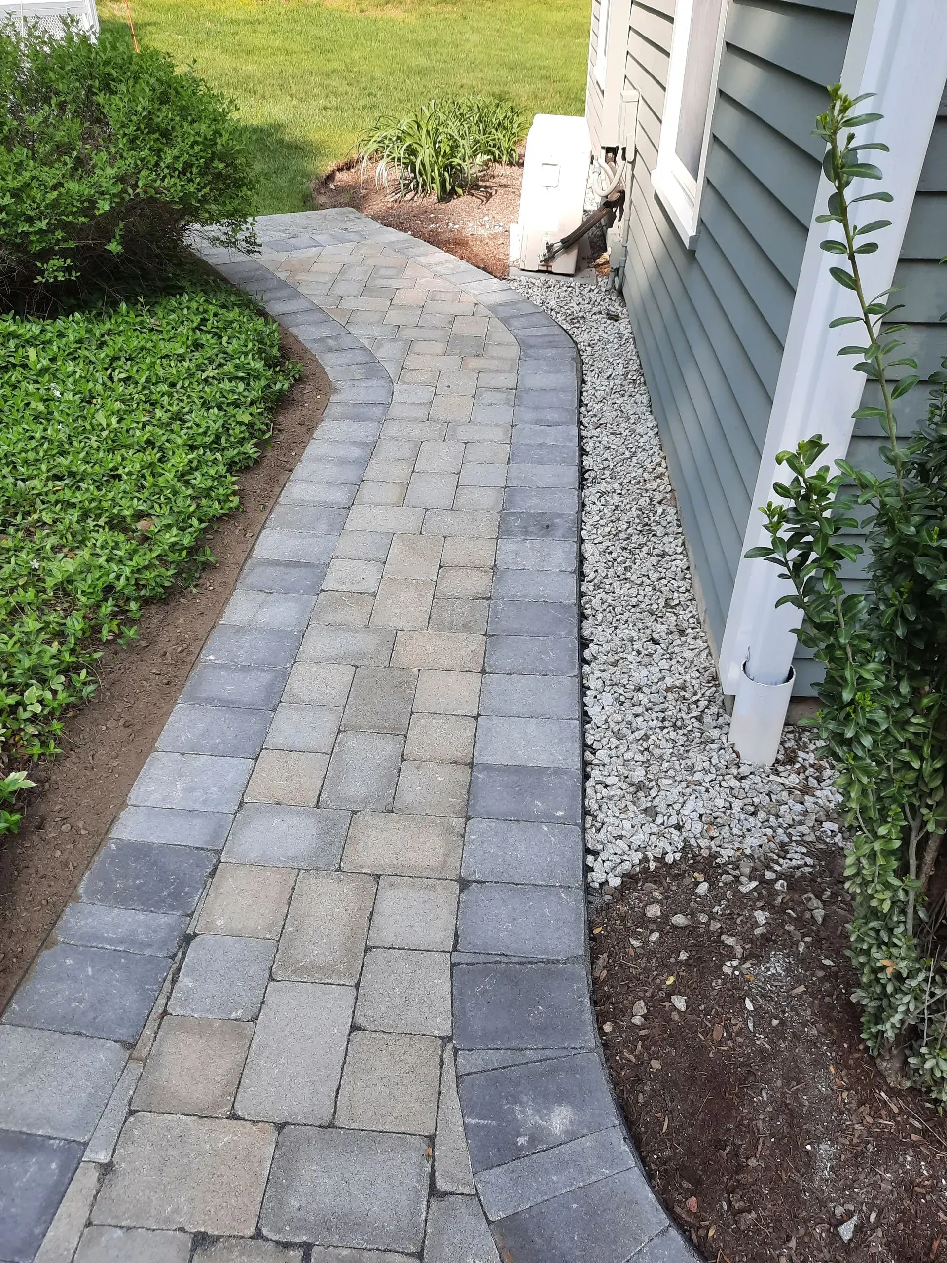 Curved gray stone walkway beside a house, bordered by gravel, mulch, and green shrubs.