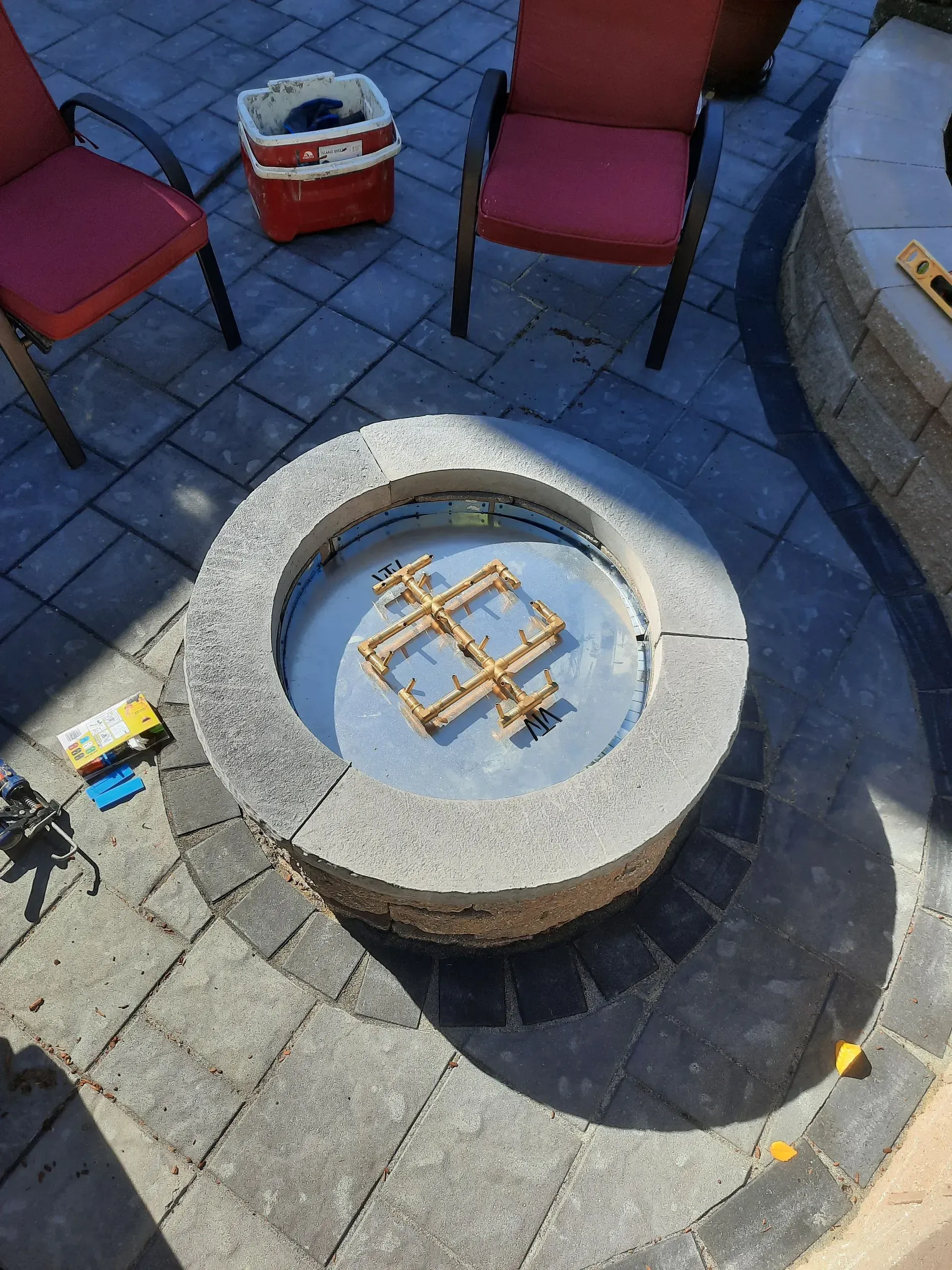 Stone fire pit in a paved patio, with red chairs and a cooler nearby.