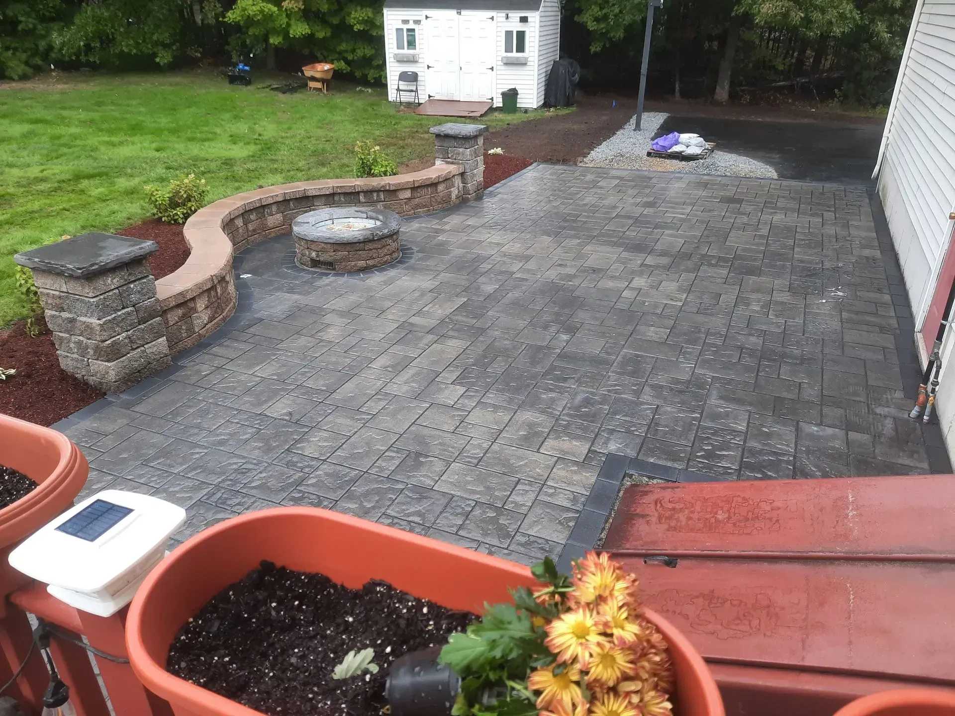Backyard patio with gray pavers, stone seating walls, and a white shed beyond a grassy lawn.