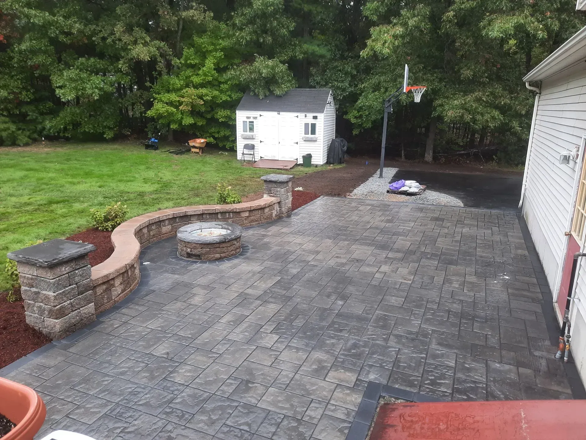 Paved backyard patio with stone seating wall, fire pit, small shed, and trees in the background