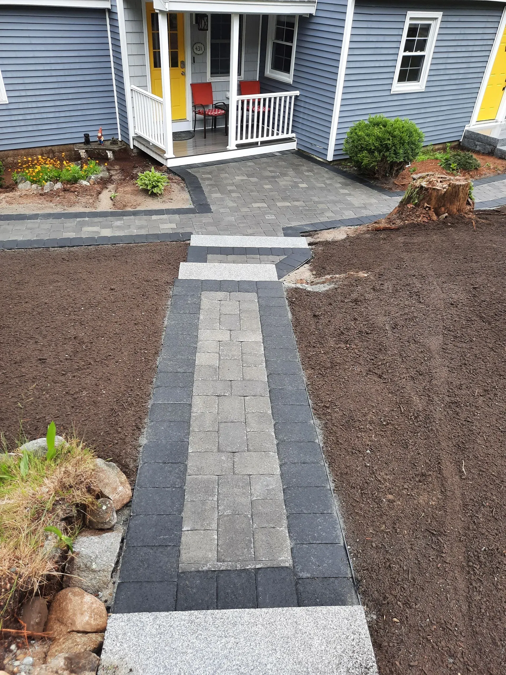 Stone walkway leading to a blue house porch with white railing and a yellow chair, bordered by gravel beds