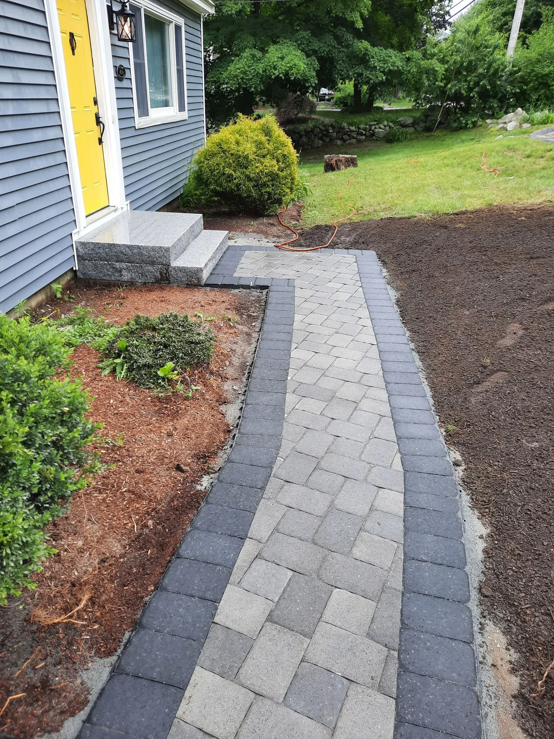 Curved gray paver walkway beside a house, bordered by mulch beds and green lawn.