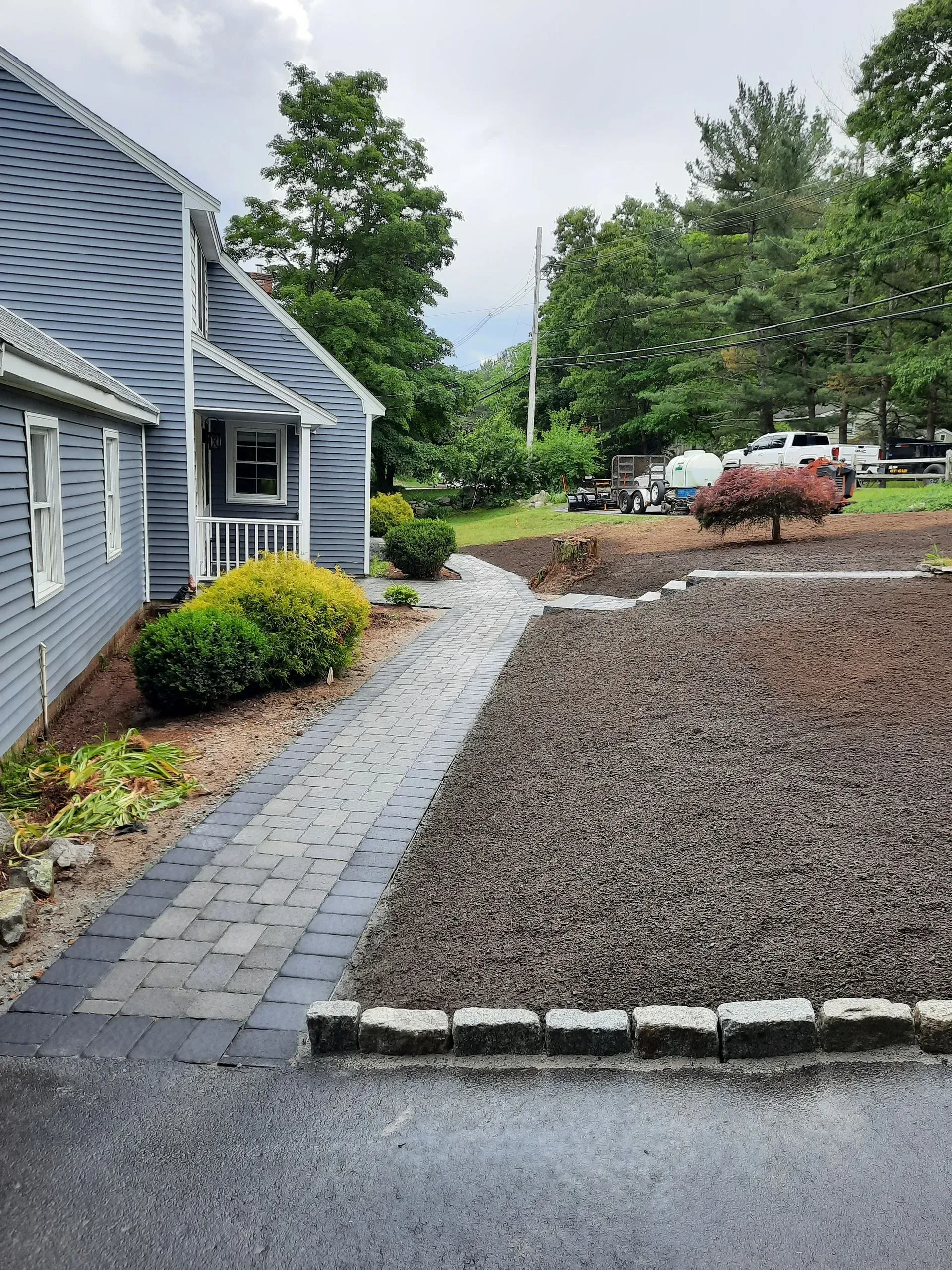 Stone walkway beside a gray house leading to a gravel parking area with shrubs and trees