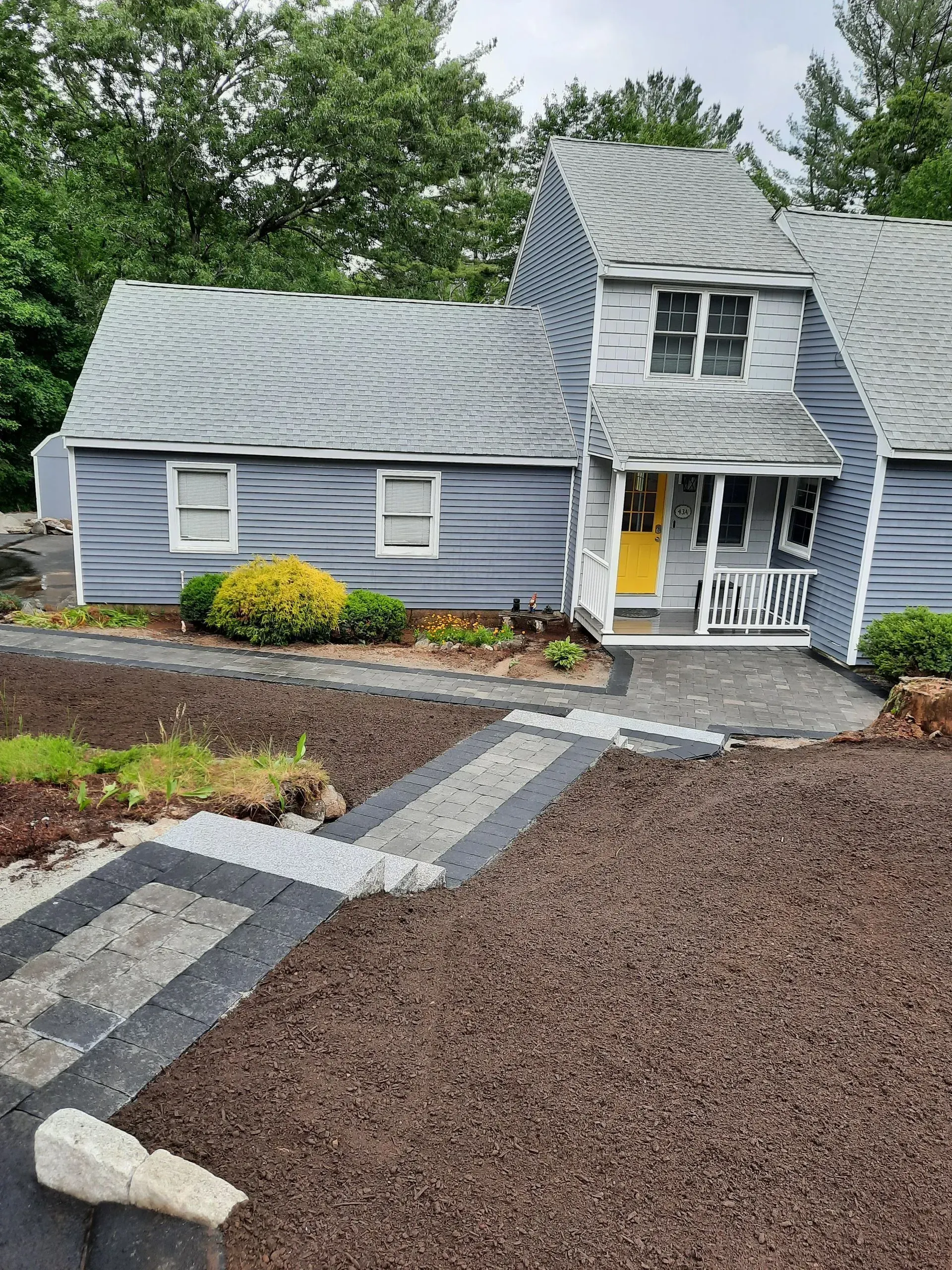 Small gray house with wavy roof, white trim, and yellow door beside a stone path and mulch garden.