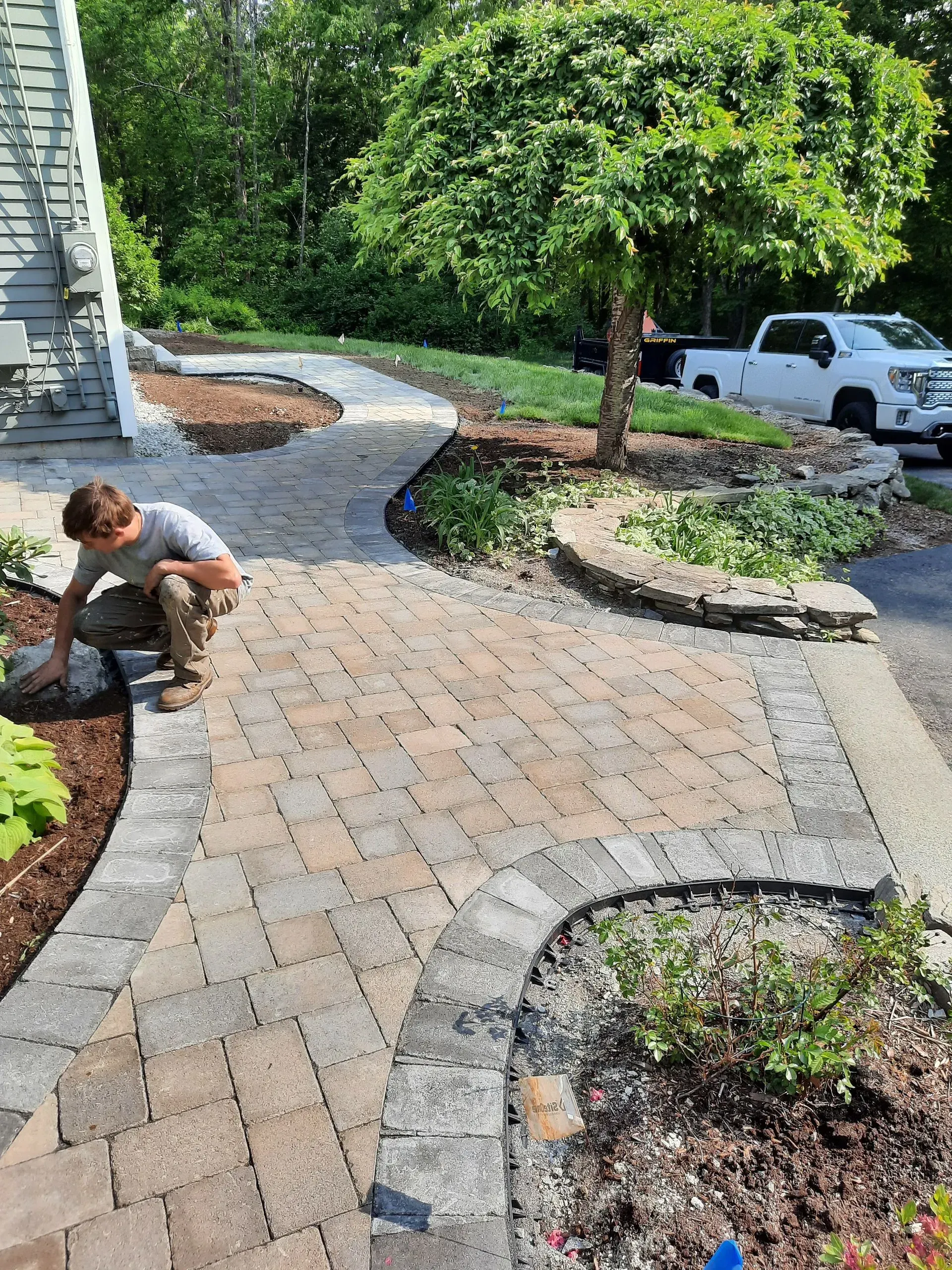 Worker installing a curved paver walkway beside a house and landscaping beds