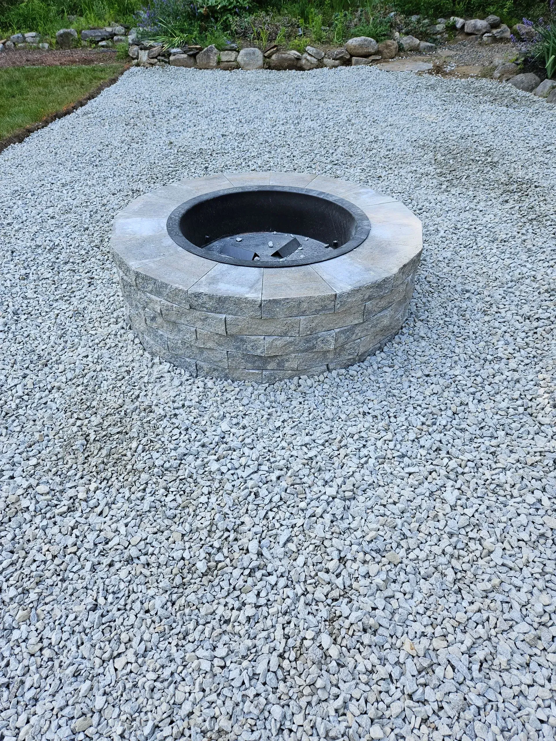 Circular stone fire pit centered in a gravel yard with grass and rocks at the edge