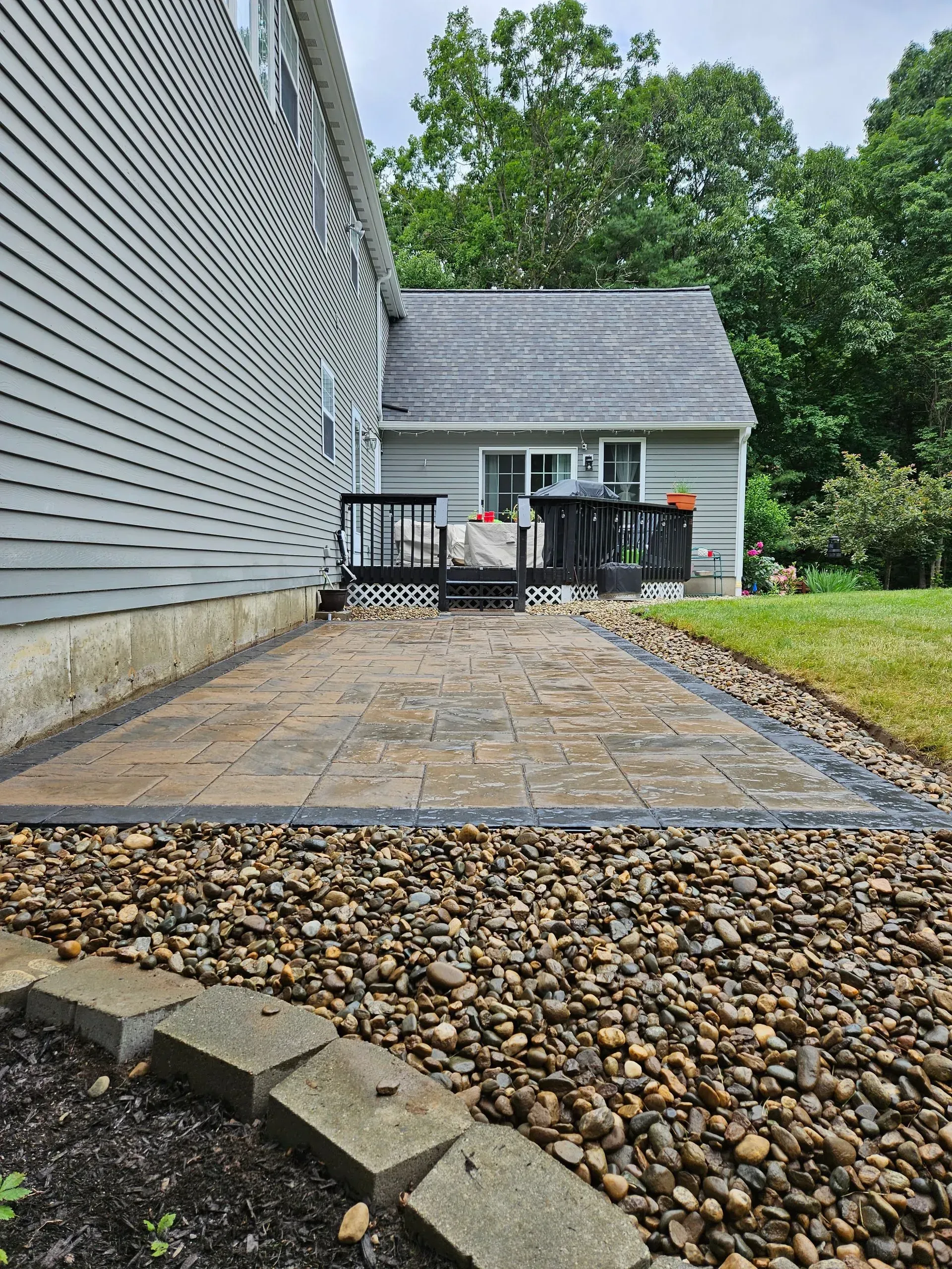Brick patio beside a gray house, with a grill, fence, and trees in a backyard.