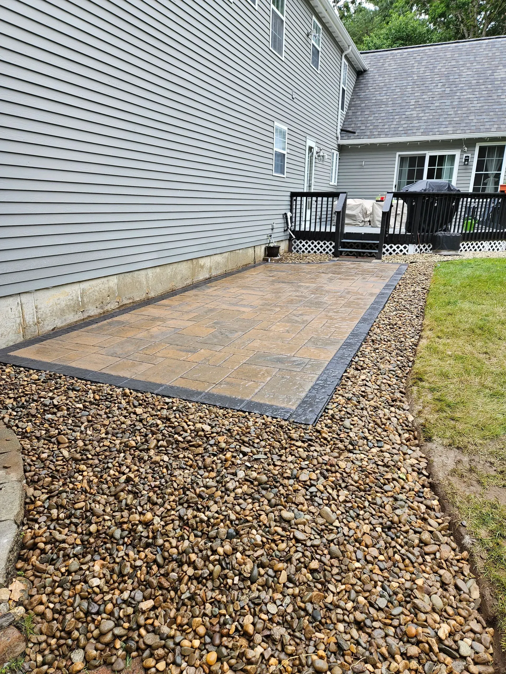 Gravel patio beside a gray house, bordered by dark edging and leading to a small deck.