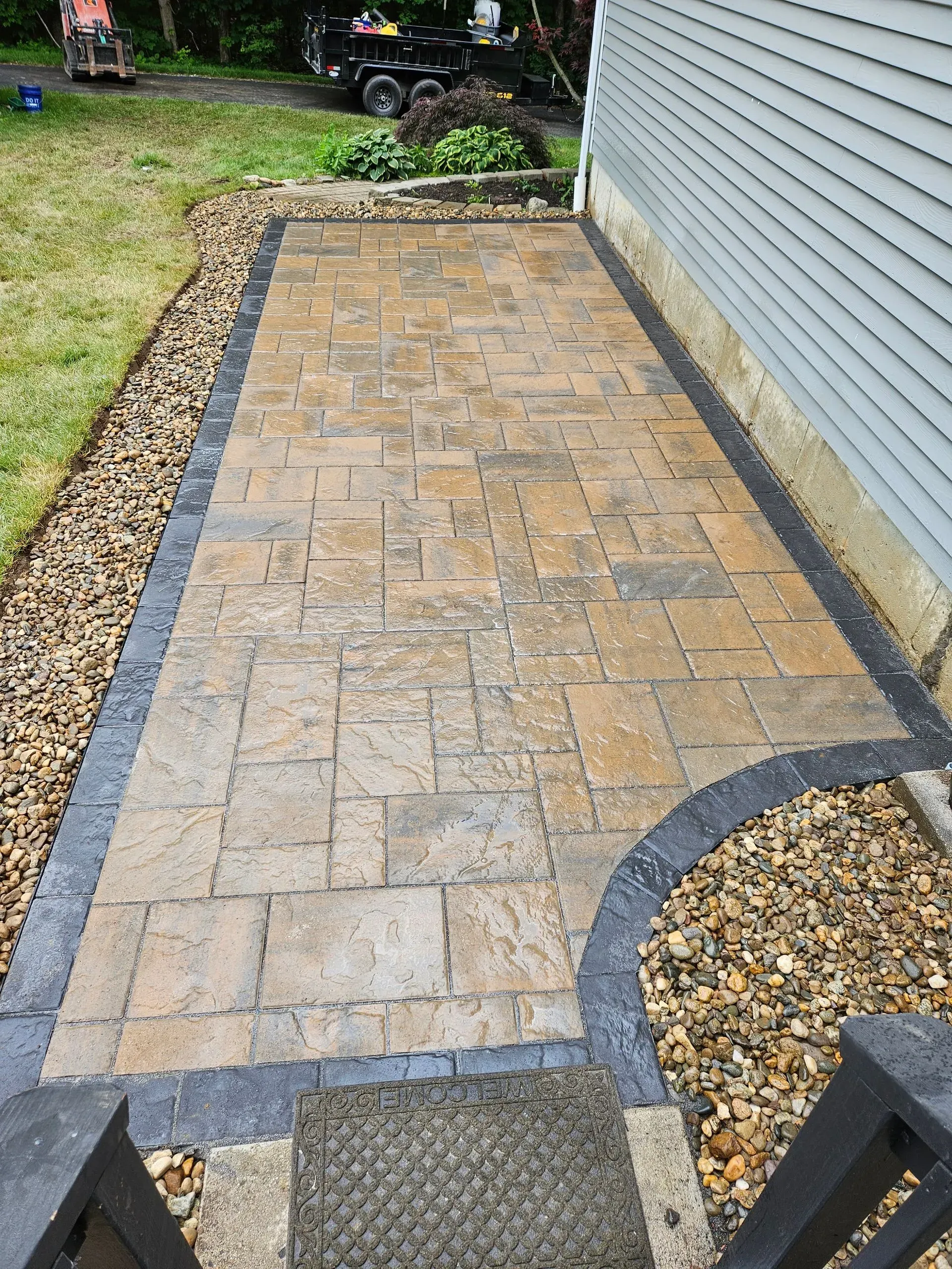 Narrow brick patio beside a house, bordered by dark edging and gravel with a curved corner.